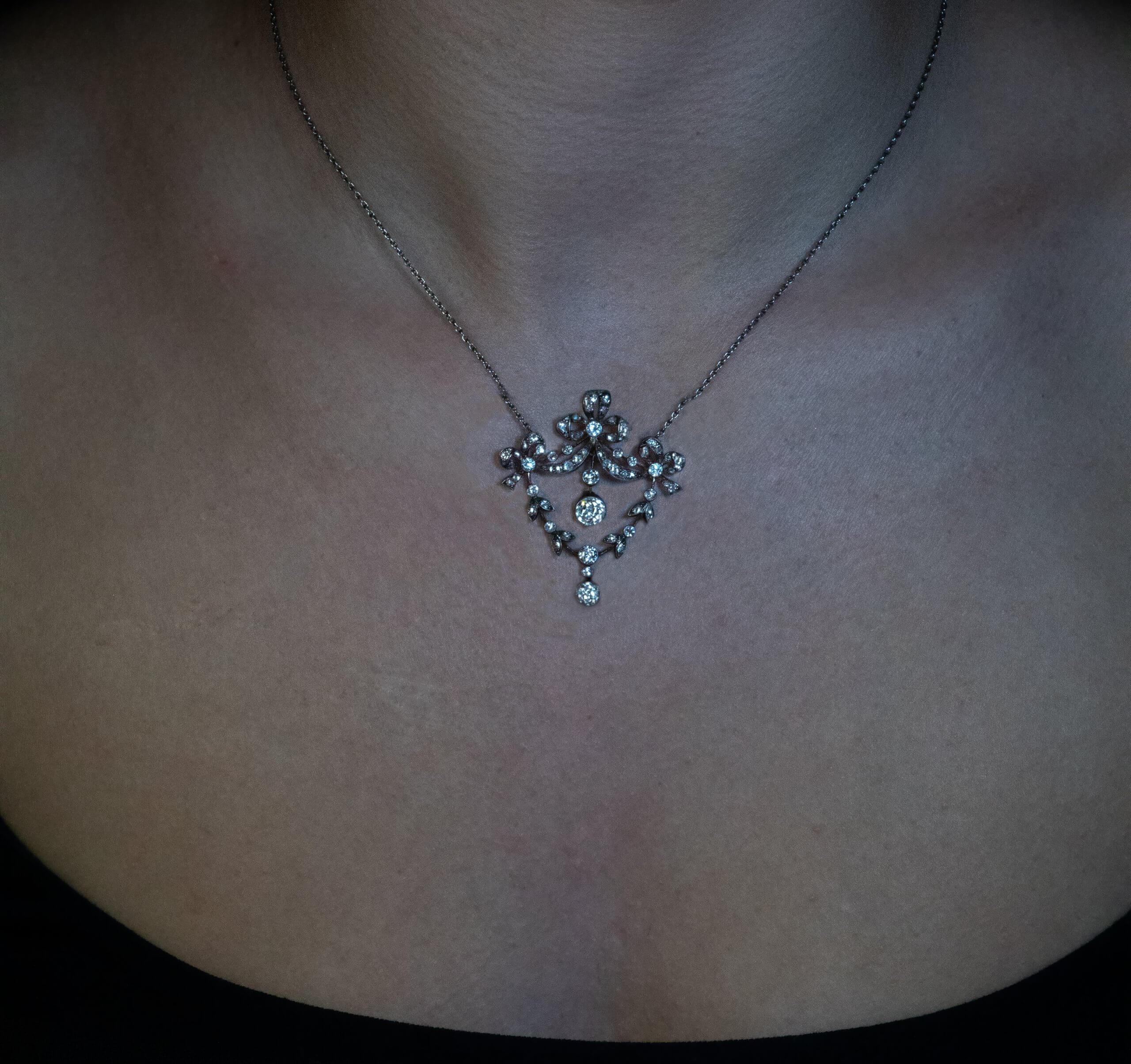 Circa 1900  This Belle Époque convertible necklace / brooch of a bow and garland motif is finely crafted in silver topped 14K gold (front – silver, back – gold) and is embellished with old mine cut diamonds.  The principal diamond measures 6.1 x 5.9