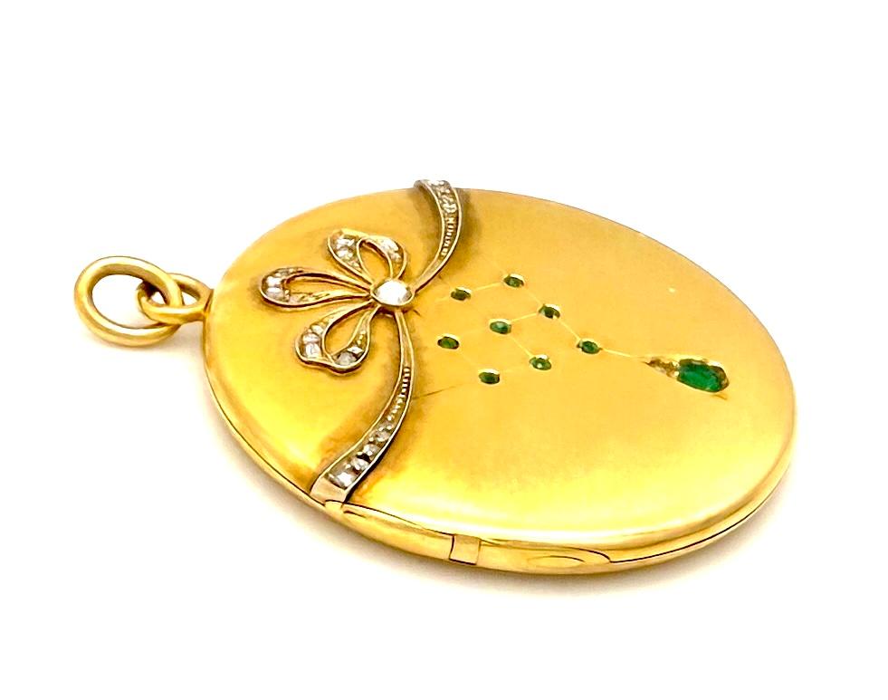 This elegant round locket was created in the last decade of the 19th century. It is created out of 18K gold and set with emeralds and rose cut diamonds. From a bow set with rose diamonds in different sizes there are engraved geometric parallel lines