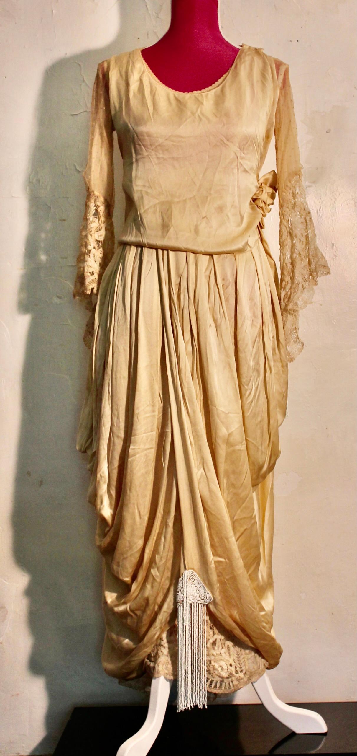 Kleid aus ecrufarbenem Seidensatin mit Brüsseler Spitze und gepunktetem Tüll an den Ärmeln sowie mit Perlenquasten an den Ärmeln und am unteren Teil des Kleides.