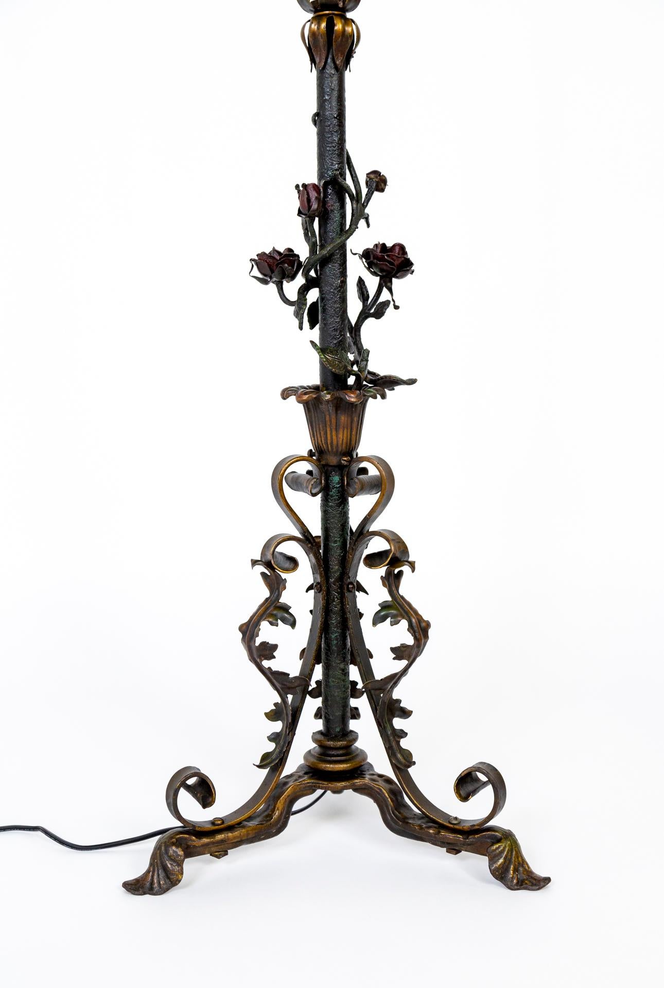A late 19th century, ornate, black, iron floor lamp with three feet narrowing into a stem adorned with roses, leaves, and scrolls; subtly tinted in red, bronze, and green. The design elements are cast and forged with precision and detail. The stem