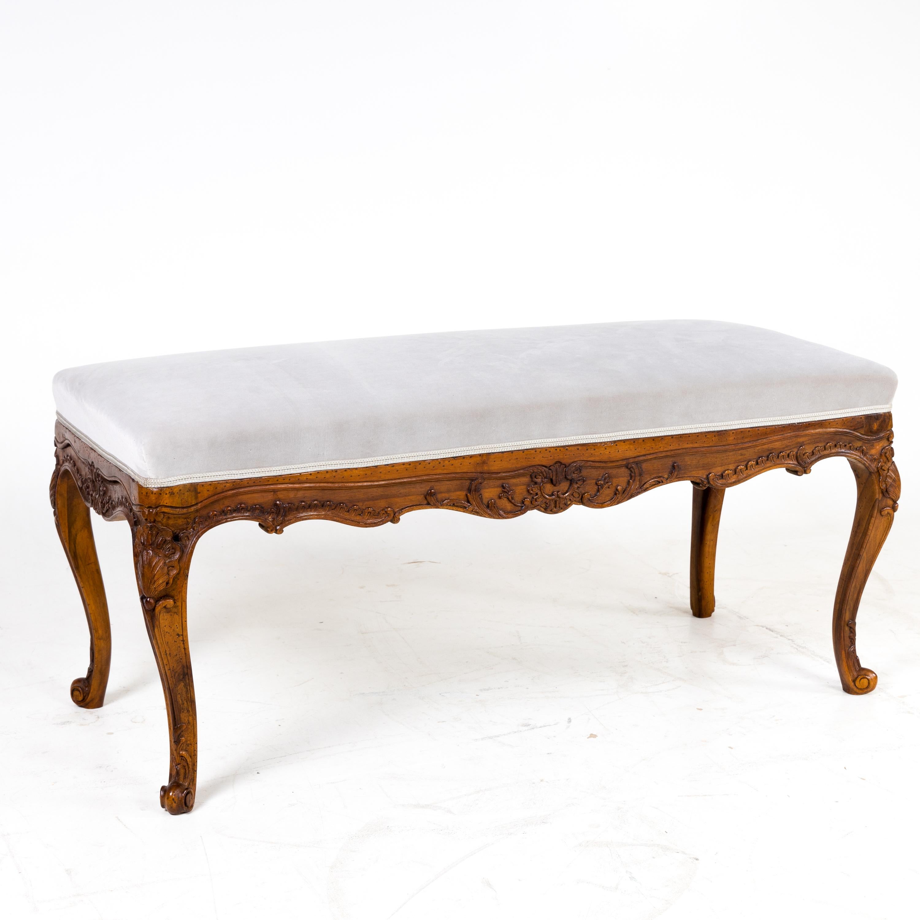 Baroque style bench standing on S-shaped curved volute feet. The frame has multiple curvatures and is decorated with carved shell and tendril decoration. The bench has been newly covered with a silver-grey velvet fabric.