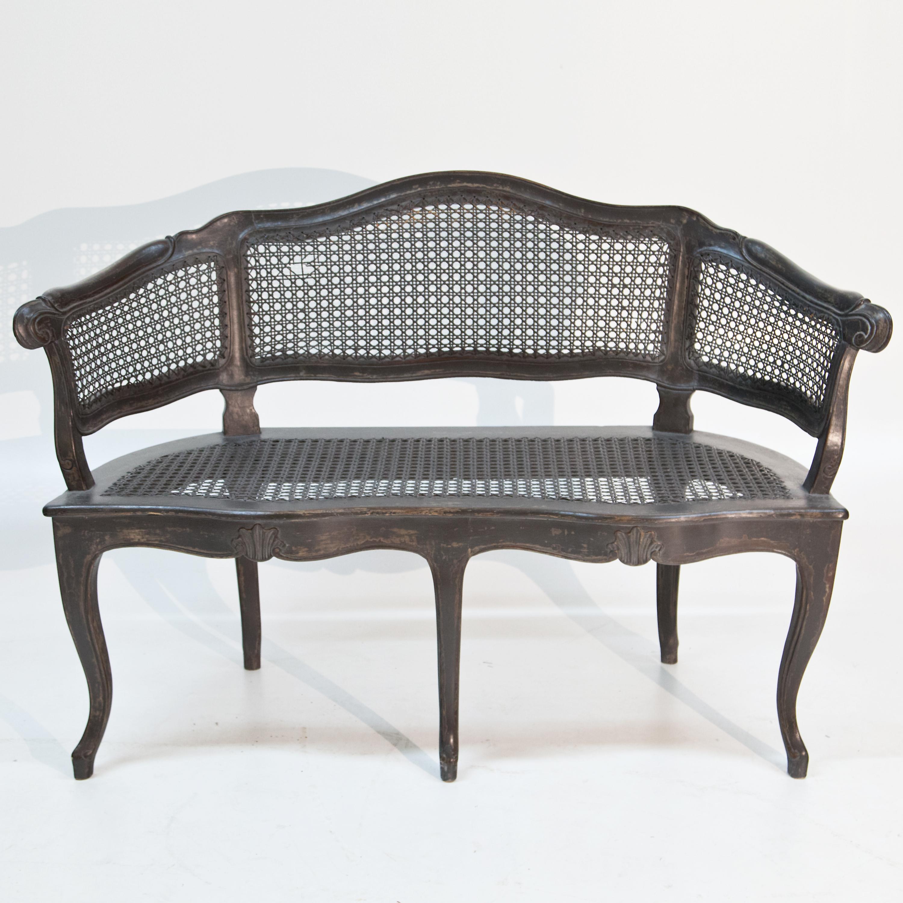 Bench with rocaille and shell decoration standing on five curved legs. The three-part backrest sinks in waves towards the sides and is covered with wickerwork like the seat. The grey setting is new and has been given an antique patina.