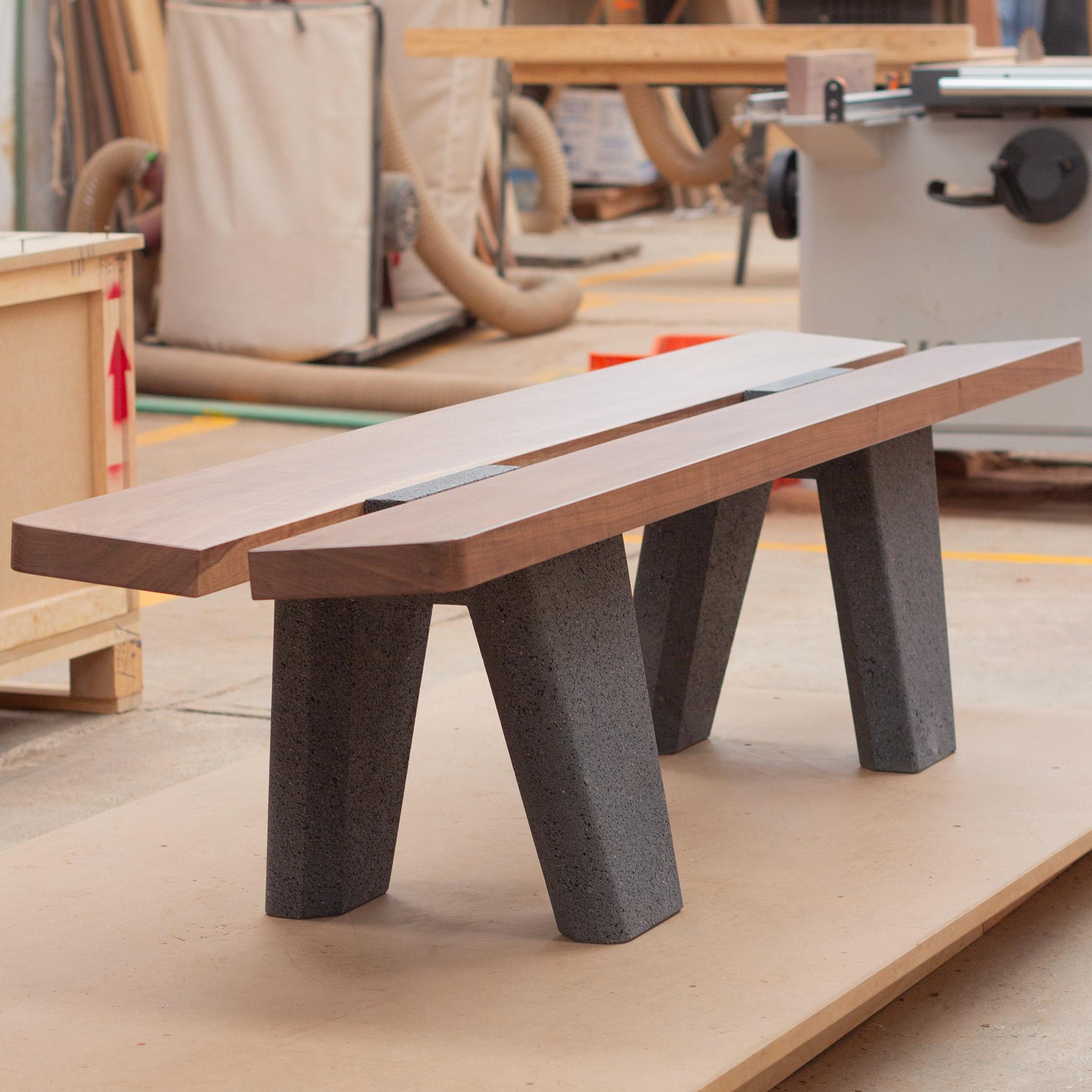 This striking bench features two robust bases sculpted from a single monolithic block of lava stone, their well-defined geometric forms evoking the grandeur of architecture. Resting atop them is a thick slab of solid wood, its rich, organic grain