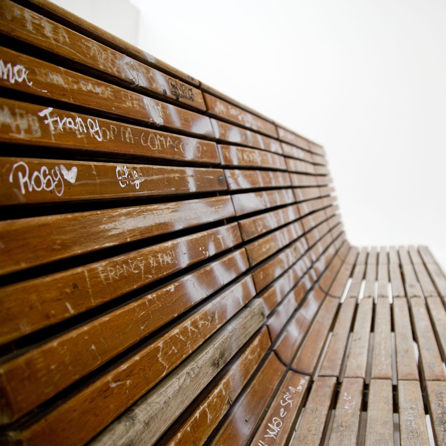 Bench on six tapered legs with horizontal bars and space for four persons. The bench shows some graffiti and is originally from an Italian train station. 12 benches are available in total.