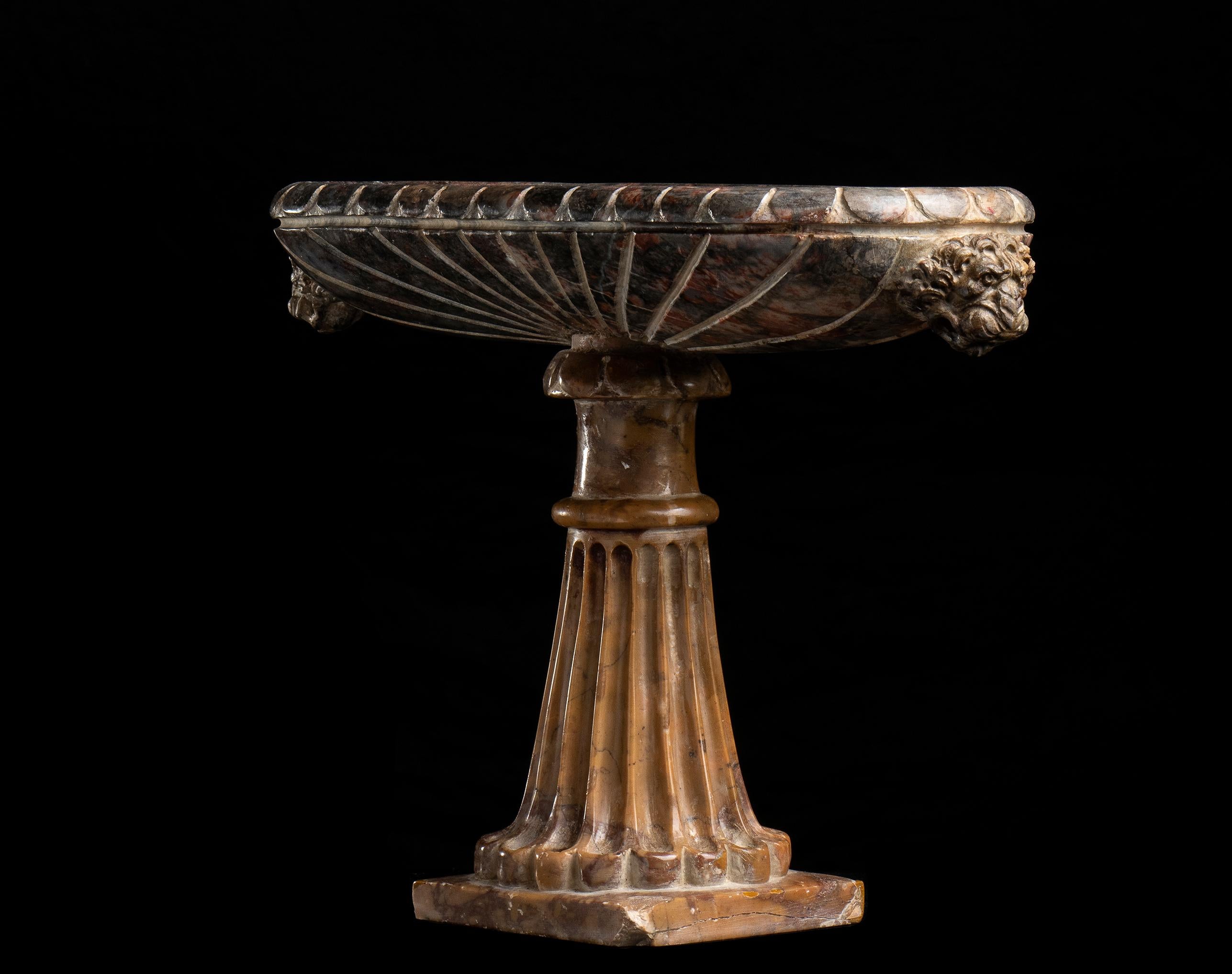 African marble stand resting on a pedestal in antique yellow marble. The elliptical basin model decorated with radial grooves from the center outwards and with pods on the upper edge and a pair of lion heads sculpted with an open mouth and with an