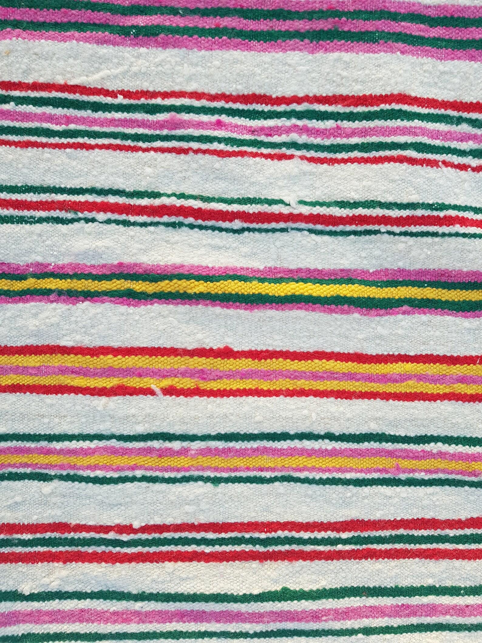 This handwoven Algerian Berber rug is a beautiful piece of craftsmanship with a simple stripped design and vibrant colors. This generous size rug features stripes of various colors such as red, yellow, green and many more all set against a beige