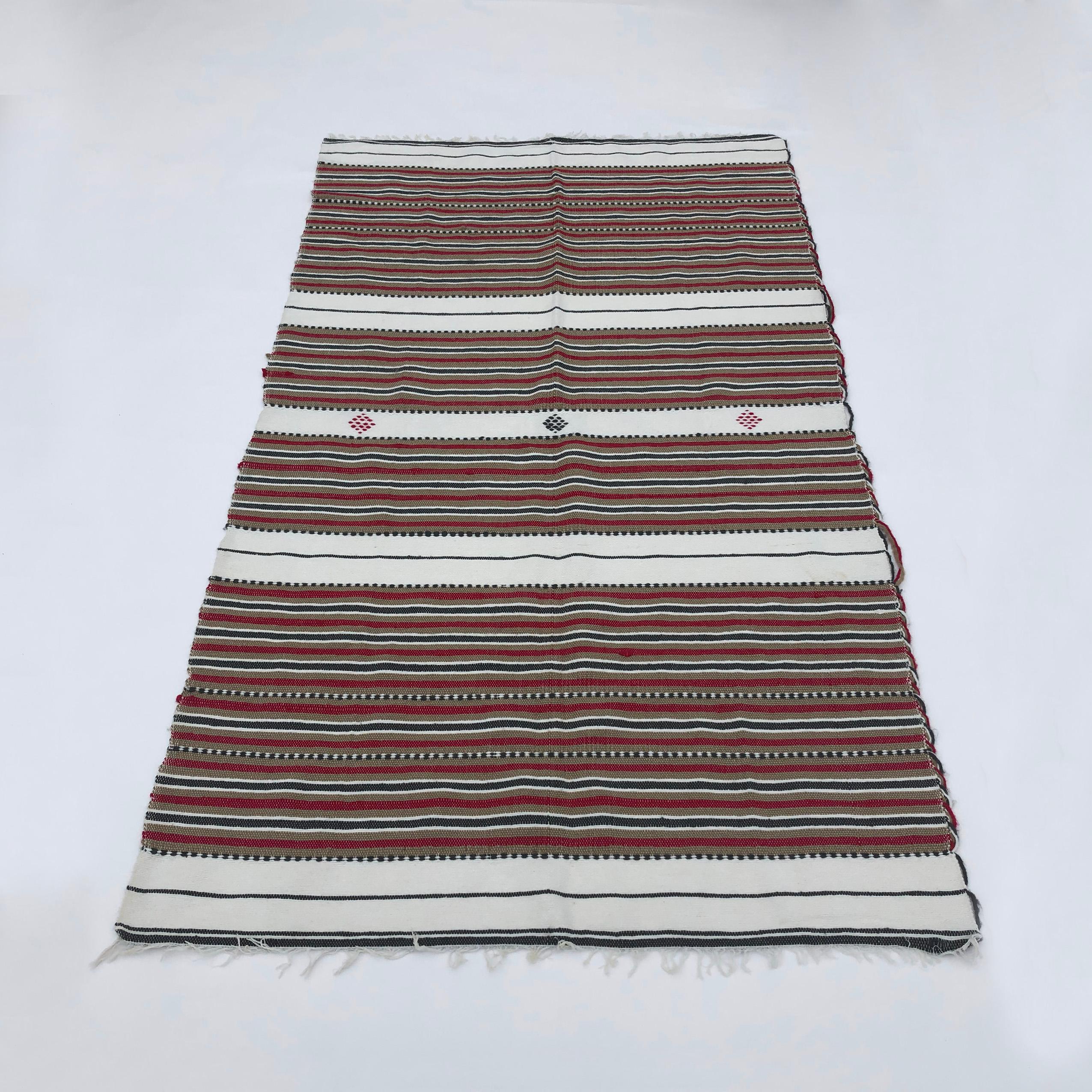 A vintage handmade wool rug from 1970s Algeria, in tricolour stripes of pleasing reds,  browns, and blacks. The wool yarns come from ethically grown livestock, and are hand dyed and treated by locals using traditional organic methods – hence the