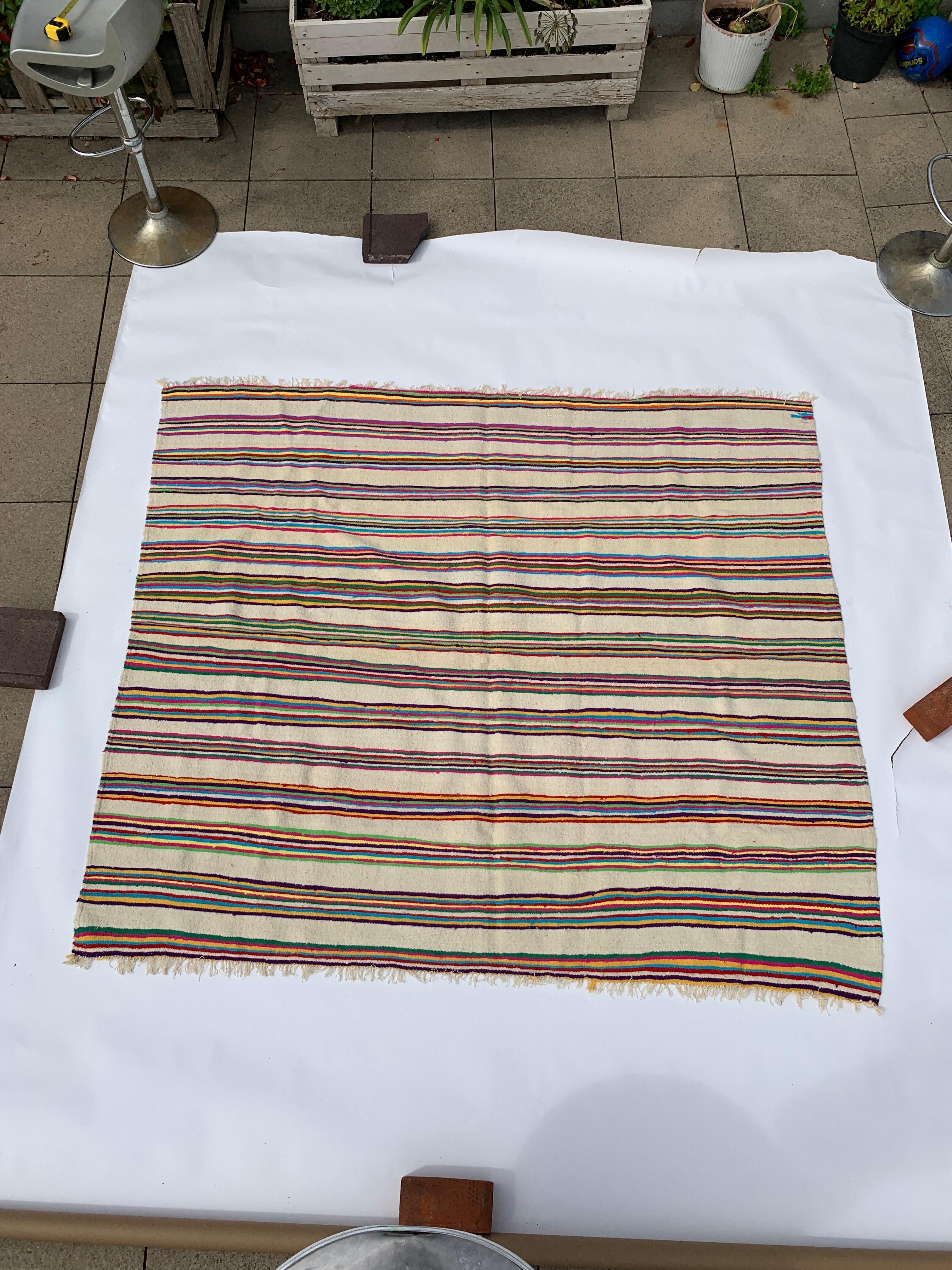 Ein Vintage-Teppich aus Berberwolle in beiger Grundfarbe mit mehrfarbigen Streifen, die in Gruppen angeordnet sind. Die Streifengruppen sind in Zehnerreihen angeordnet. 

Die mehrfarbigen Streifen des Teppichs sind visuell auffällig und schaffen ein
