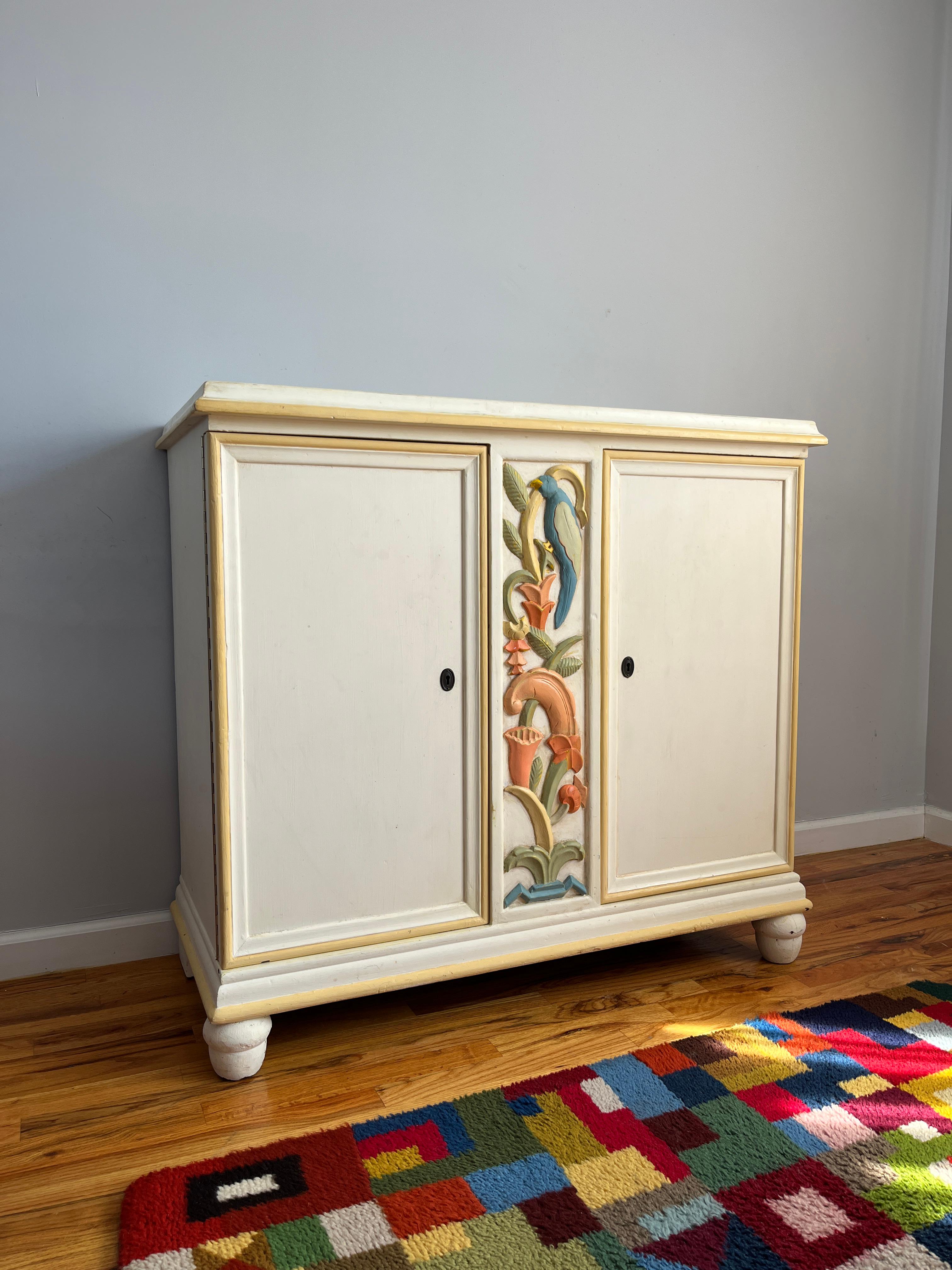 Cette armoire à deux portes présente un panneau central sculpté de motifs floraux et aviaires stylisés en relief polychrome. Le meuble est peint en ivoire avec des reflets jaunes le long des moulures. A l'intérieur se trouve une étagère réglable.