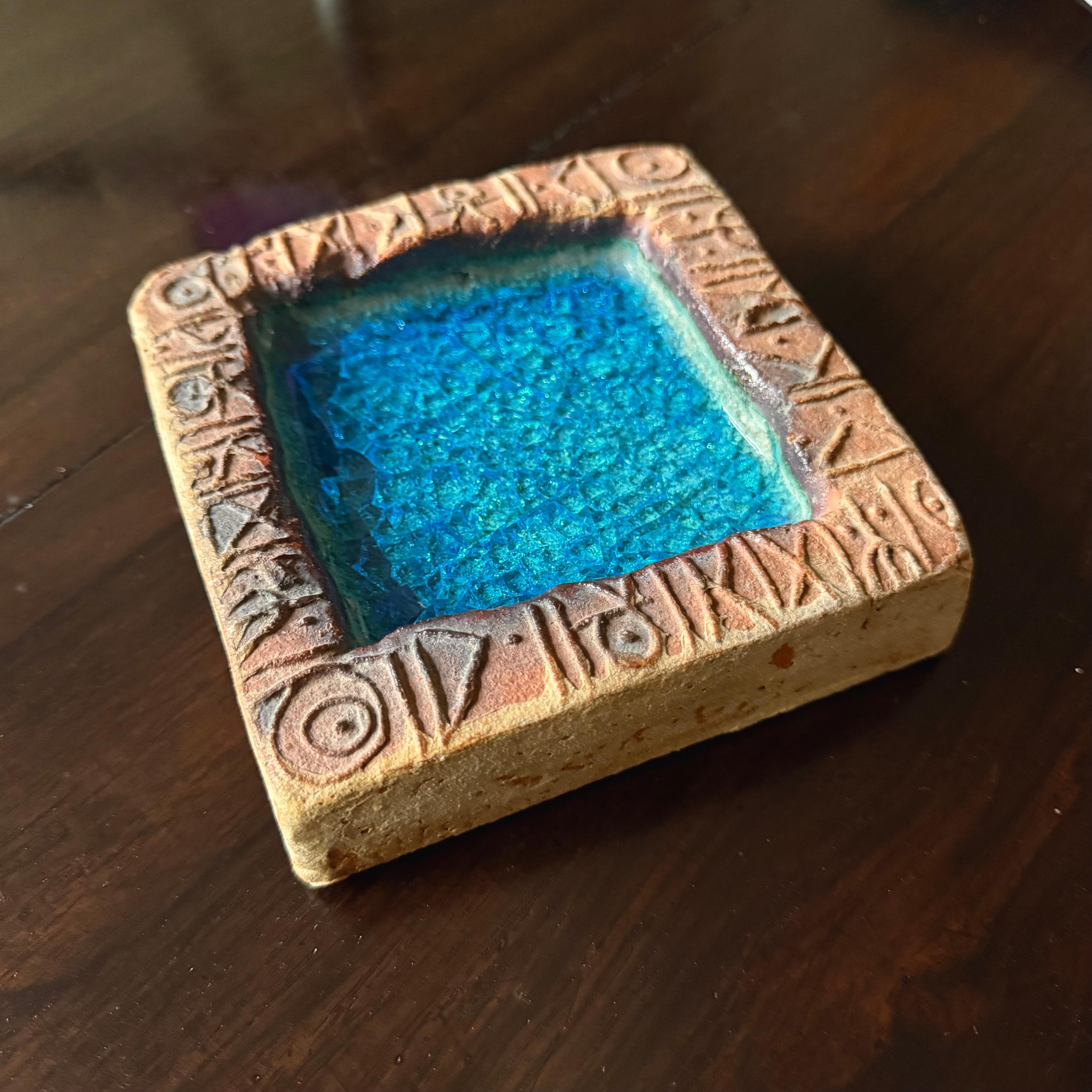 Un piatto decorativo o vide-poche del ceramista Bernard Rooke. Inghilterra, metà del XX secolo. Ceramica fatta a mano, in toni naturali; la base in vetro blu con finitura crepata. La decorazione a rune di Rooke corre intorno alla parte superiore e,
