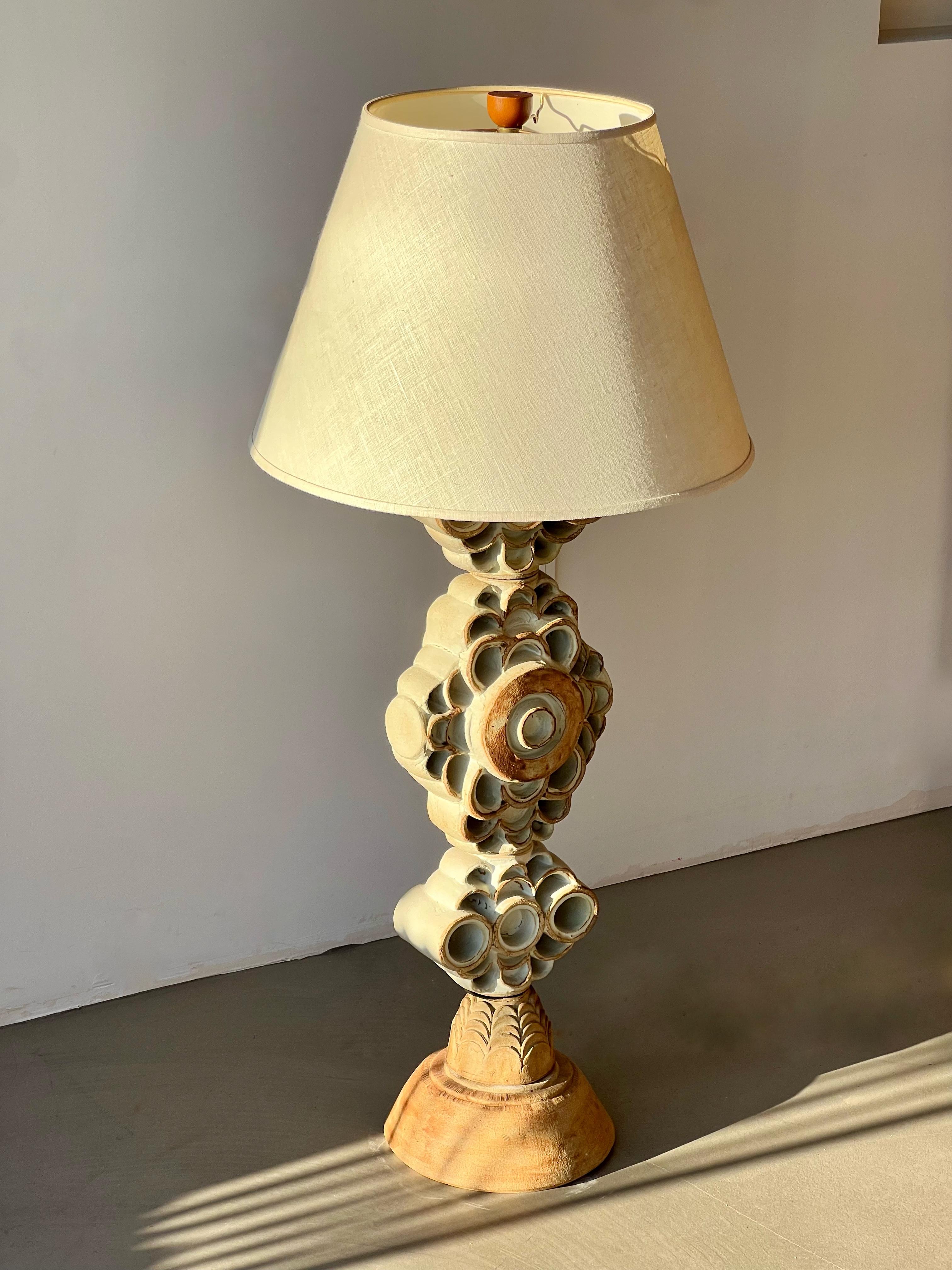 The lamp features a sculptural ceramic base adorned with intricate, textured patterns and earthy tones that evoke a sense of warmth and natural beauty. Its stacked, modular design is both striking and versatile, making it an ideal focal point for