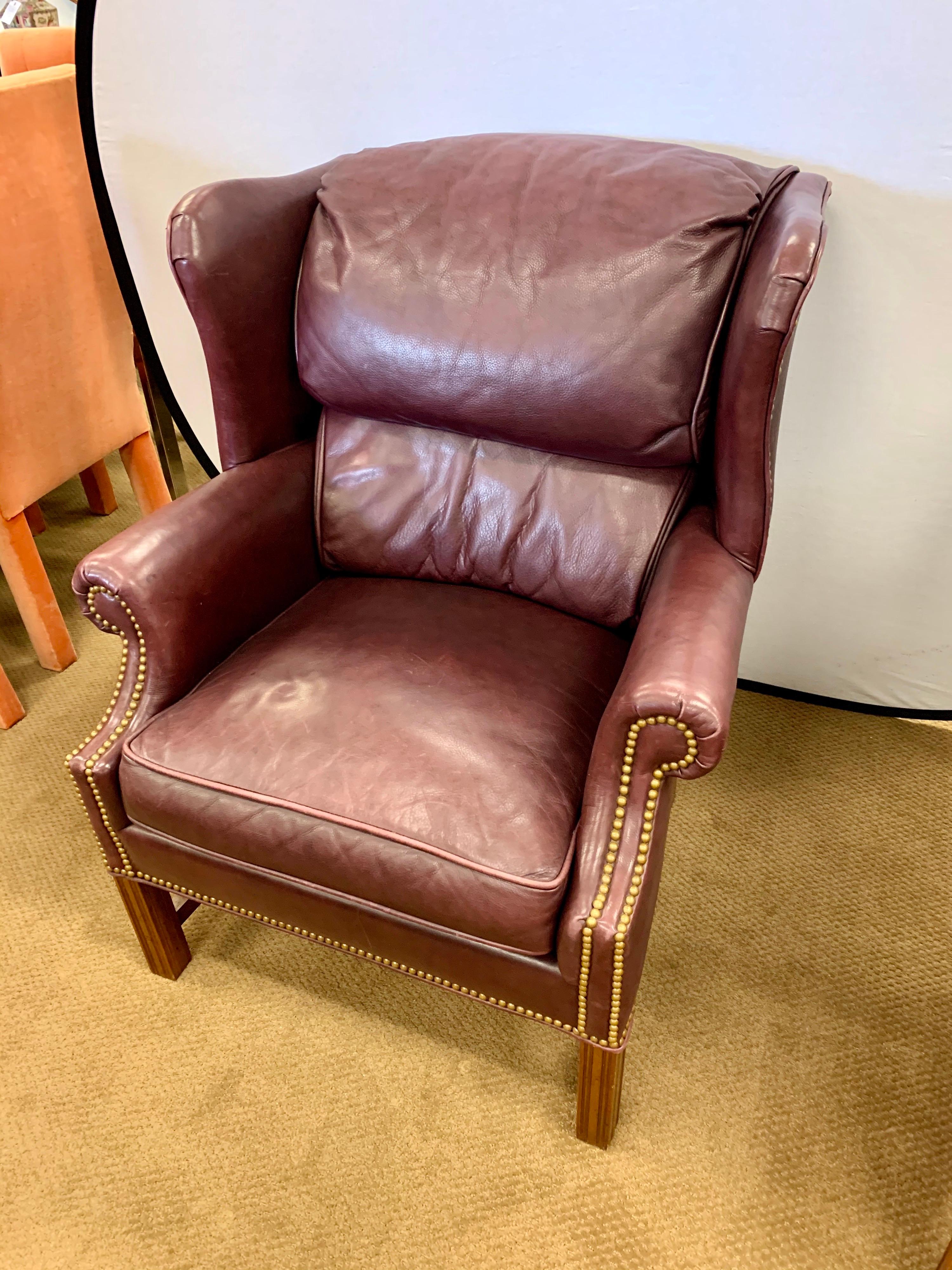 Grand, welcoming and wonderful leather wingback club chair from the late 20th century, by Bernhardt. In the manner of George III. Having mahogany legs with carvings throughout. Gorgeous merlot leather and brass nailhead accents. This elegant