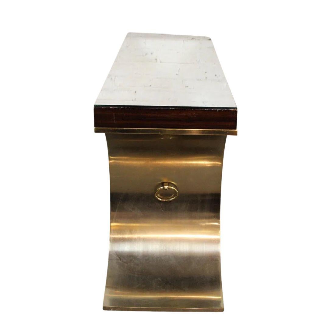 Karl Springer style console table featuring a mirrored top supported by sculptural, shaped brass base with a ring on each side and center stretcher. The polished brass frame adds a luxurious, reflective warmth, while the mirrored surface enhances