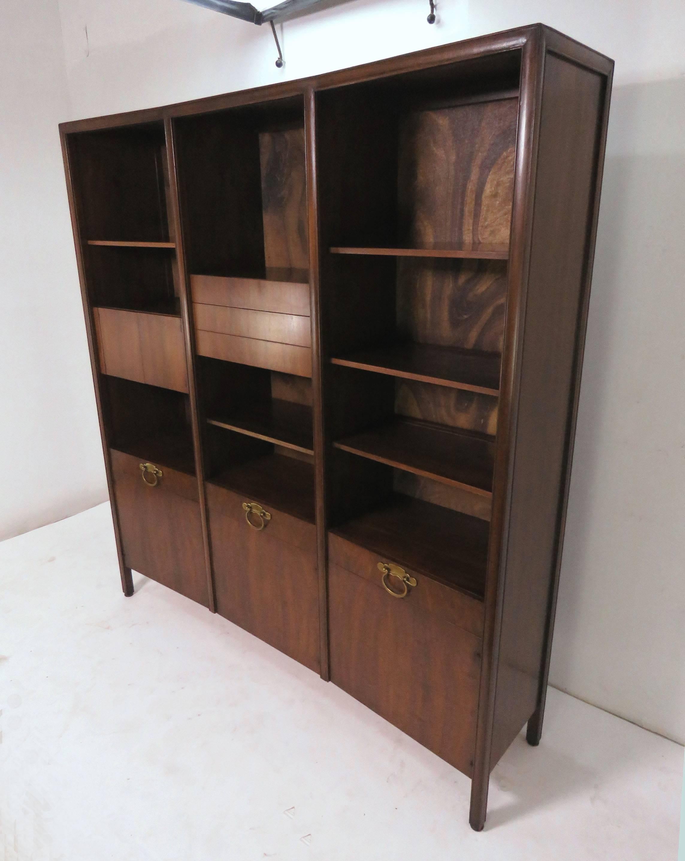 Freestanding wall unit cabinet in walnut with solid brass hardware, by Bert England for John Widdicomb. The upper cabinets and shelves are height adjustable within their three bays. The back of the unit is also finished so it may serve as a room