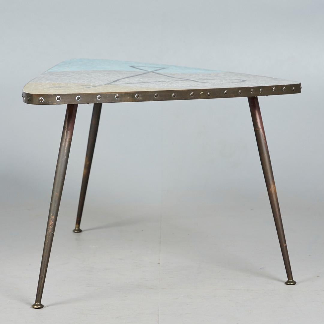 La impresionante mesa de centro de mosaico y latón diseñada por Berthold Müller-Oerlinghausen en la década de 1950 es una joya que fusiona la artesanía excepcional con un diseño vanguardista. Originaria de Alemania, esta mesa en forma de boomerang