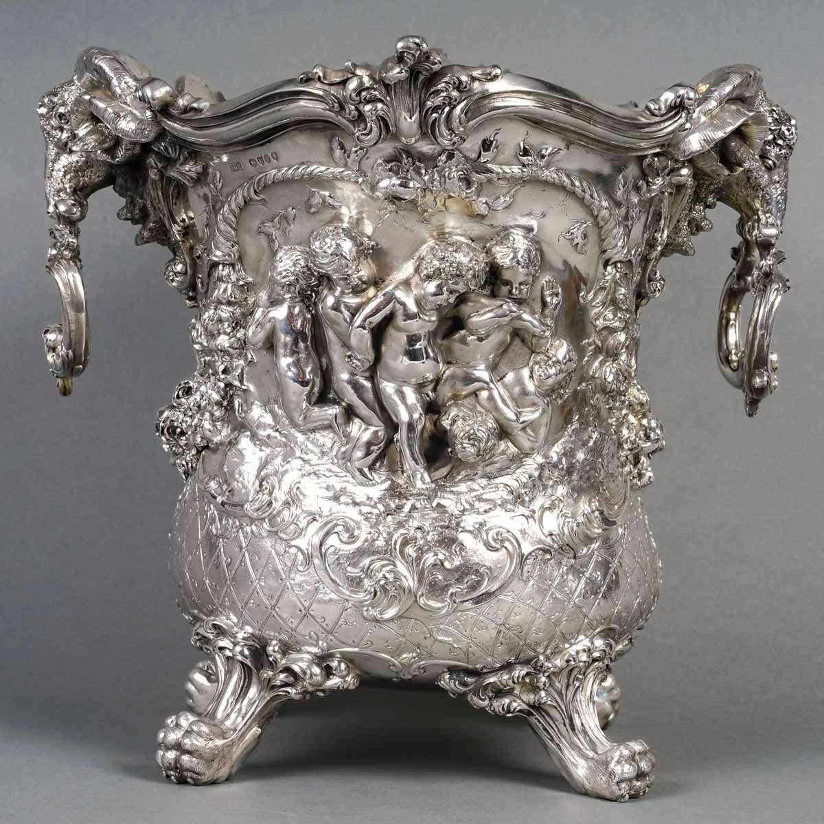 Solid silver champagne bucket in scalloped shape resting on four claw feet, Decorated in strong relief of playing putti, inserted in cartridges, surrounded by garlands of flowers and cornucopias.

The leafy handles are held by two mouflon heads.