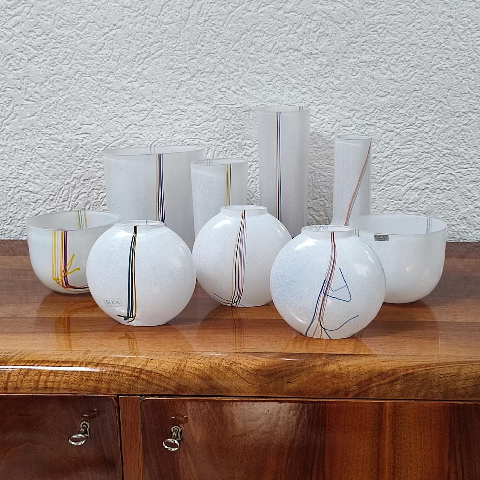 Questa collezione di nove vasi in vetro di Bertil Vallien per Kosta Boda, Svezia, risalente agli anni '80, presenta ciotole e vasi in vetro bianco con fili di vetro multicolore ai lati. Questi pezzi sono una testimonianza dell'approccio innovativo