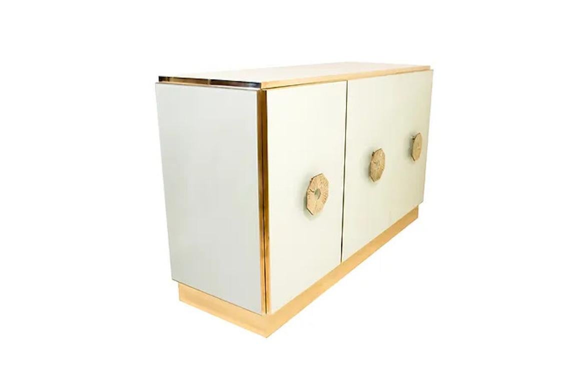 Bespoke customizable 3-door console credenza/cabinet entirely handcrafted in Italy with elegant Art Deco style and Hollywood Regency glam, the surround decorated with art glass in an elegant soft ivory cream, raised on a plinth clad in brass, the