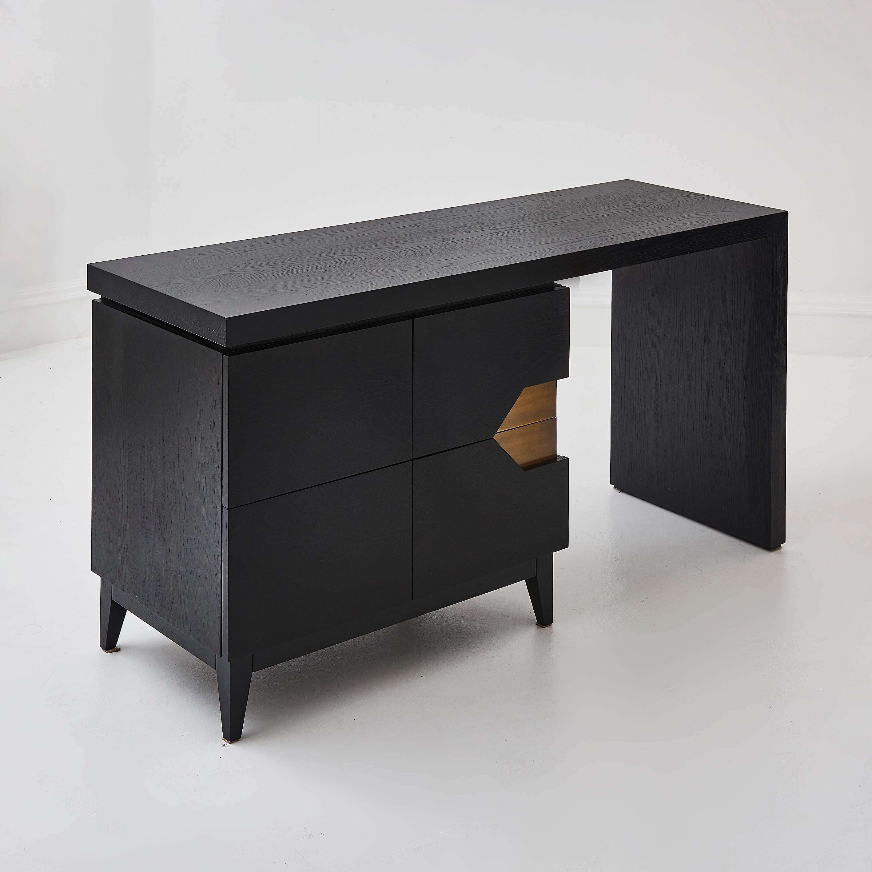 A bespoke handmade desk in a dark black ash solid wood material, with solid brass detailing on the drawers. Featuring a sleek design, this desk has four drawers in total, with two featuring a cut out brass design. 

Made from solid wood and handmade