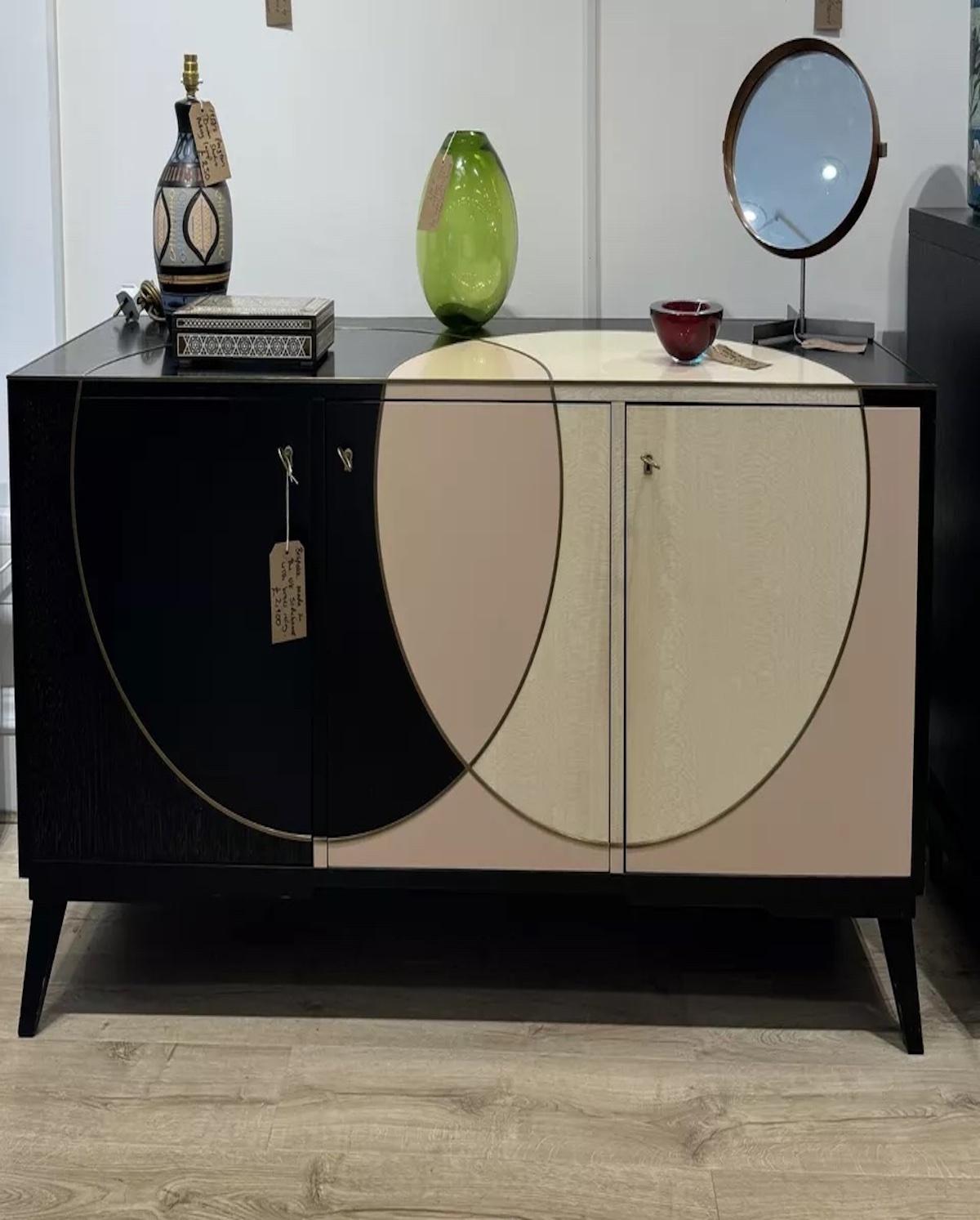 Una credenza in legno massiccio realizzata su misura nel Regno Unito, con dettagli in ottone intarsiati e un design circolare che si estende sul piano e sulle ante della credenza in un legno color rosa chiaro e crema. 

Con una serratura e una