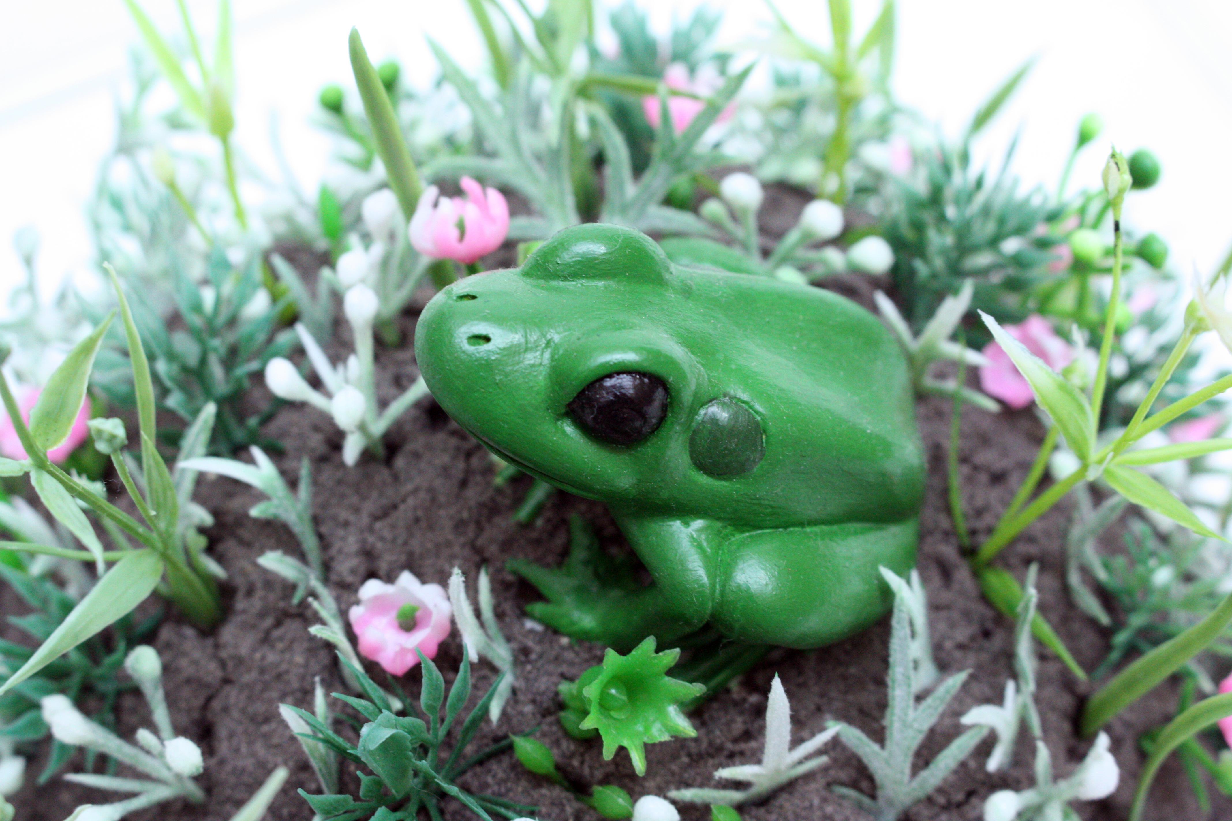 A lone ceramic frog perches atop a dirt mound covered in blooming artificial flowers.  She is a beautiful captive inside her glass aquarium and she is the last of her kind...A creature in need of protection and adoration who raises questions about