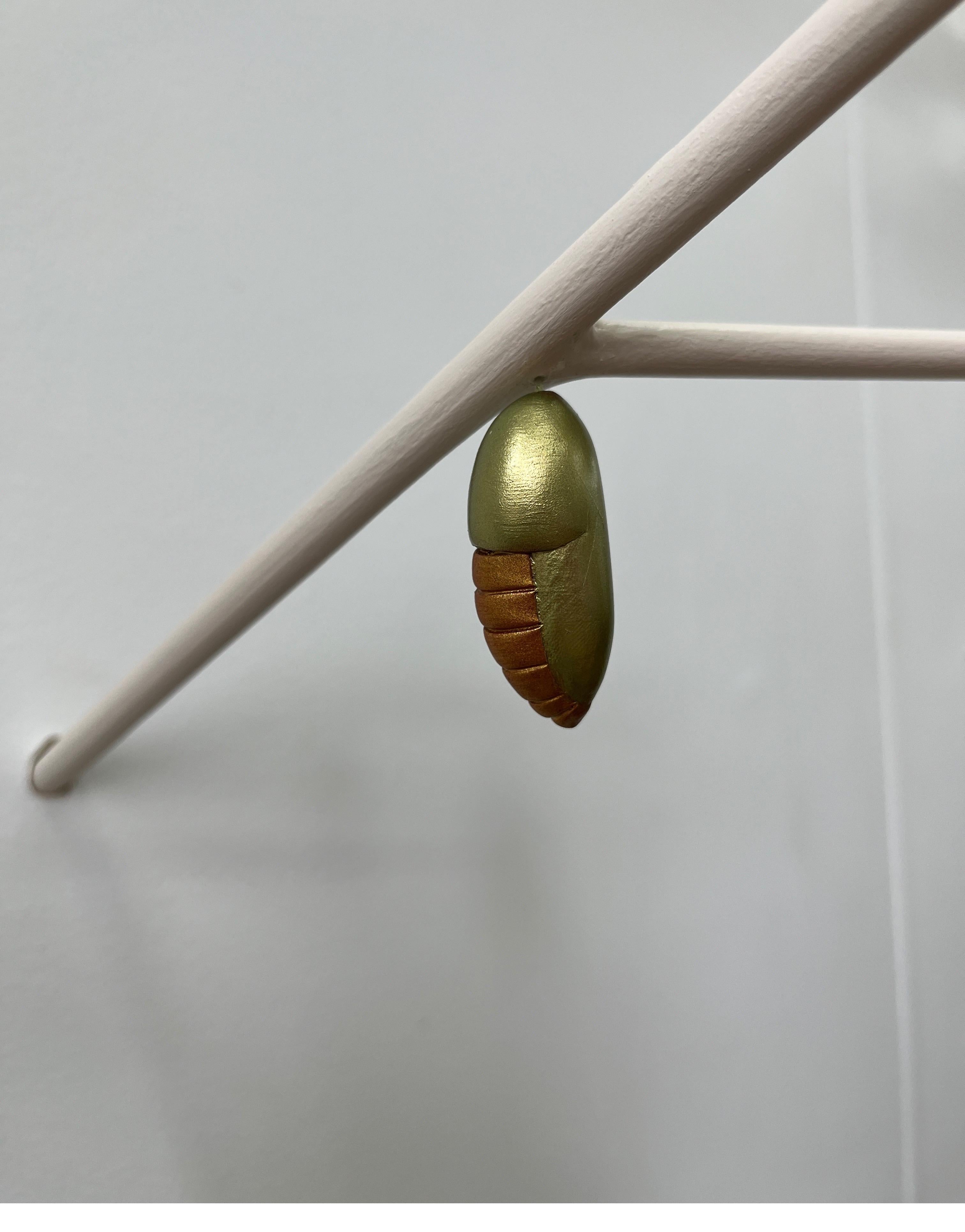 A beautiful chrysalis meticulously hand-sculpted in epoxy clay and painted with high quality metallic paint by New York artist Bethany Krull, hangs delicately from a minimalist branch.  The intimate piece extends itself from the wall into the space