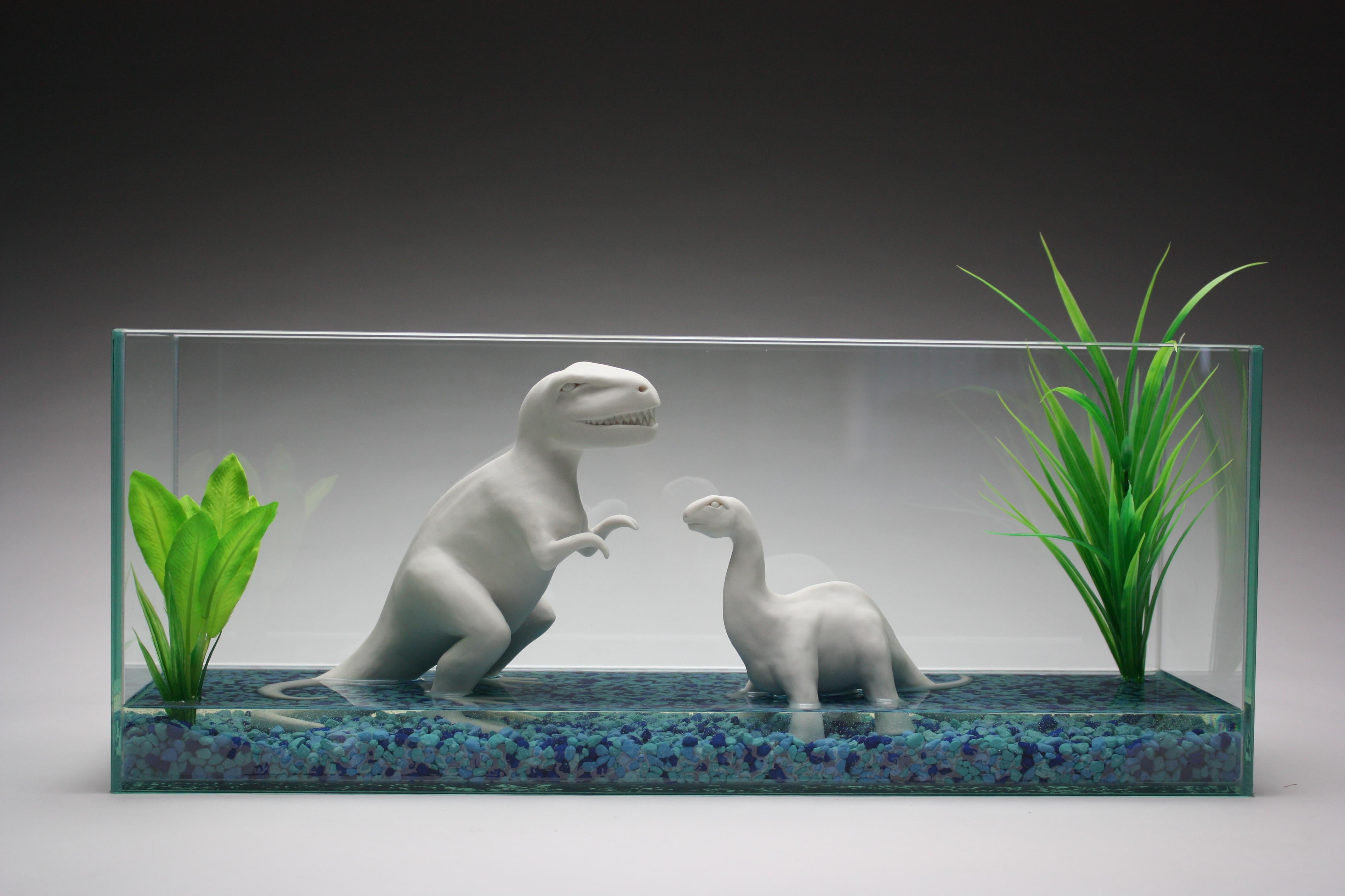 Une paire de dinosaures en porcelaine blanche fabriqués à la main par l'artiste new-yorkaise Bethany Krull sont contenus dans un aquarium en verre.  Les dinosaures, les plantes artificielles d'un vert éclatant et les pierres d'aquarium bleues sont