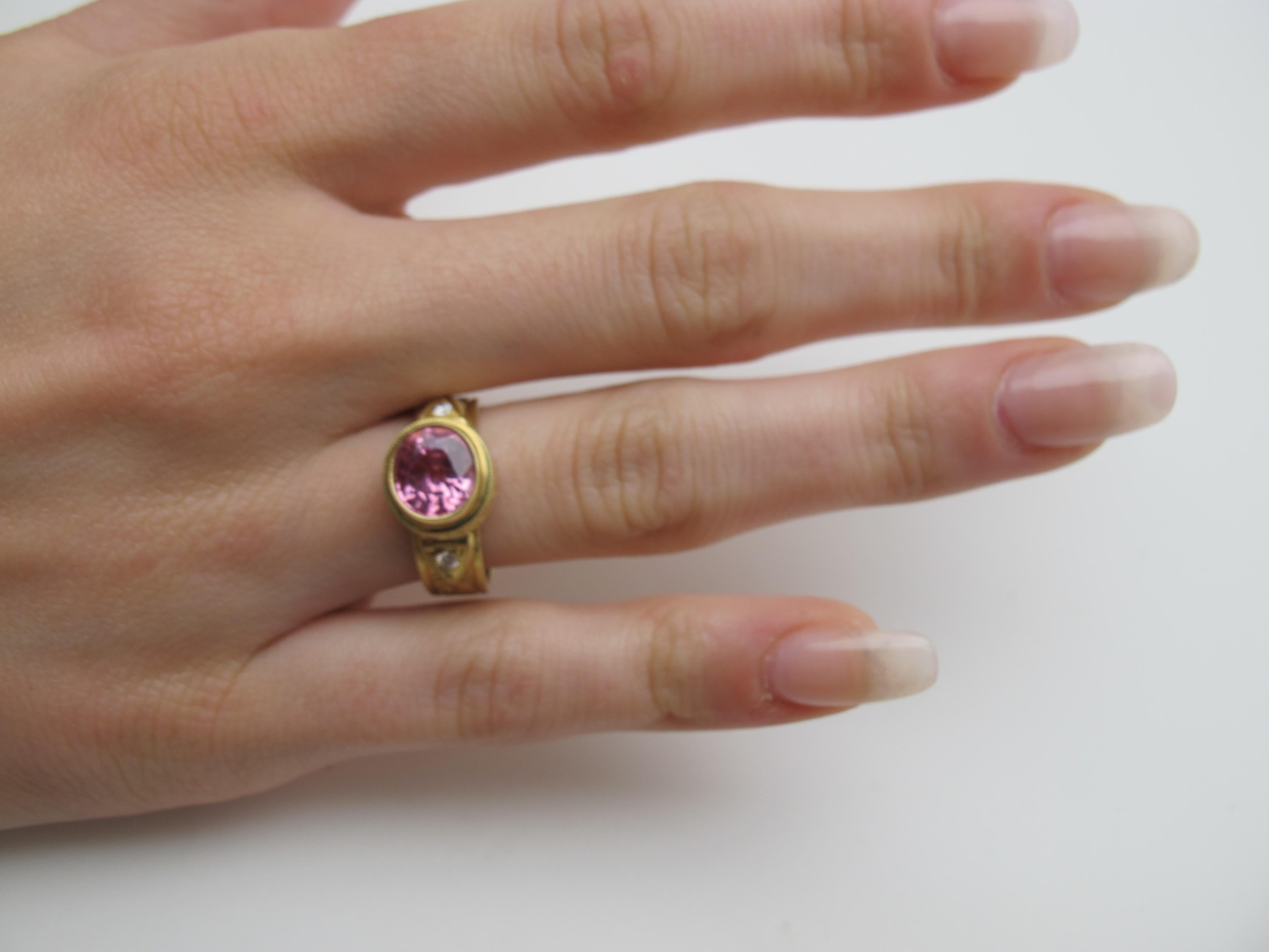 A sparkly, bright pink spinel is featured in this pretty ring.  It is oval, measuring 9x7mm and weighing 2.28 carats. It is bezel set in a beautifully handmade and hand engraved ring made of 18 karat yellow gold. Two round brilliant cut diamonds