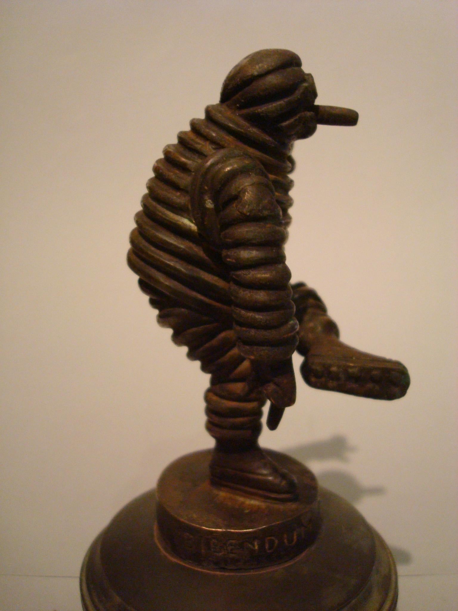 This is a highly collectible Bibendum Michelin Man bronze car mascot, Hood Ornament, Mascotte voiture.
It´s approximate 4 inch tall depiction of the Michelin man, also known as Bibendum, joyfully raises his arm while jauntily kicking up his leg.