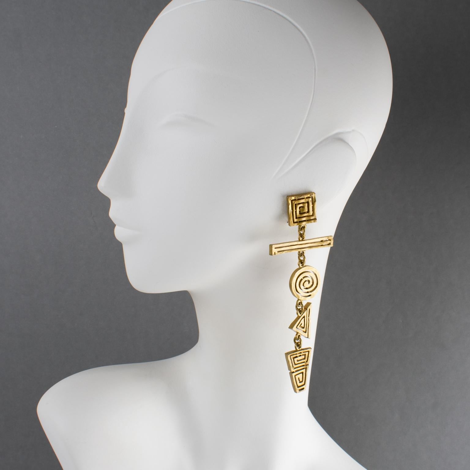 Questi rari orecchini a clip in metallo dorato dei primi anni '90 di Nelly Biche de Bere, Paris, presentano una forma extra-lunga con elementi geometrici intagliati e trasparenti. I pezzi hanno una consistenza semi-materica con una patina oro