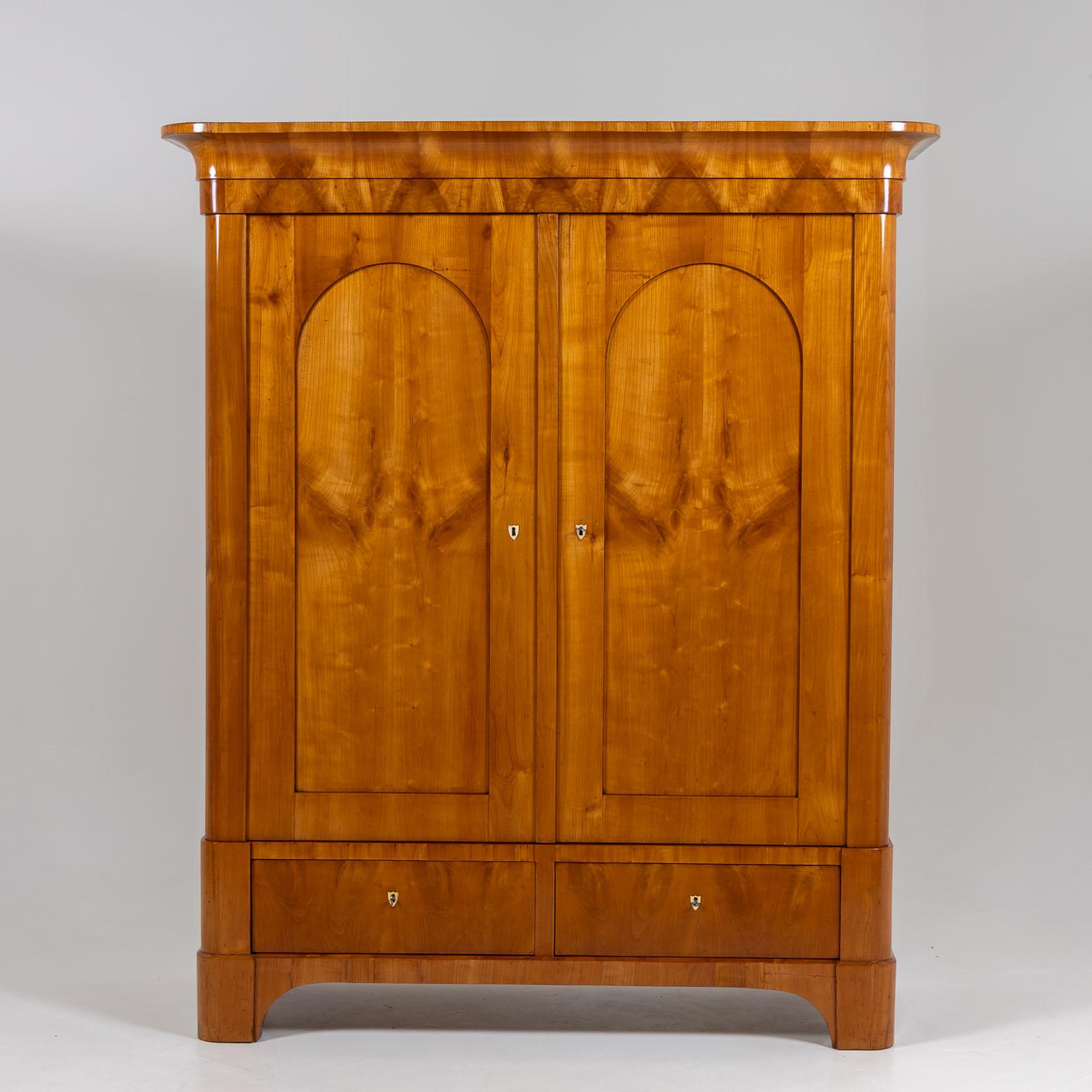 Biedermeier cabinet with two doors, two drawers and a protruding cornice. The wardrobe is veneered in cherry and has been polished by hand. The doors have panels with segmental arches. The interior is fitted with three shelves.