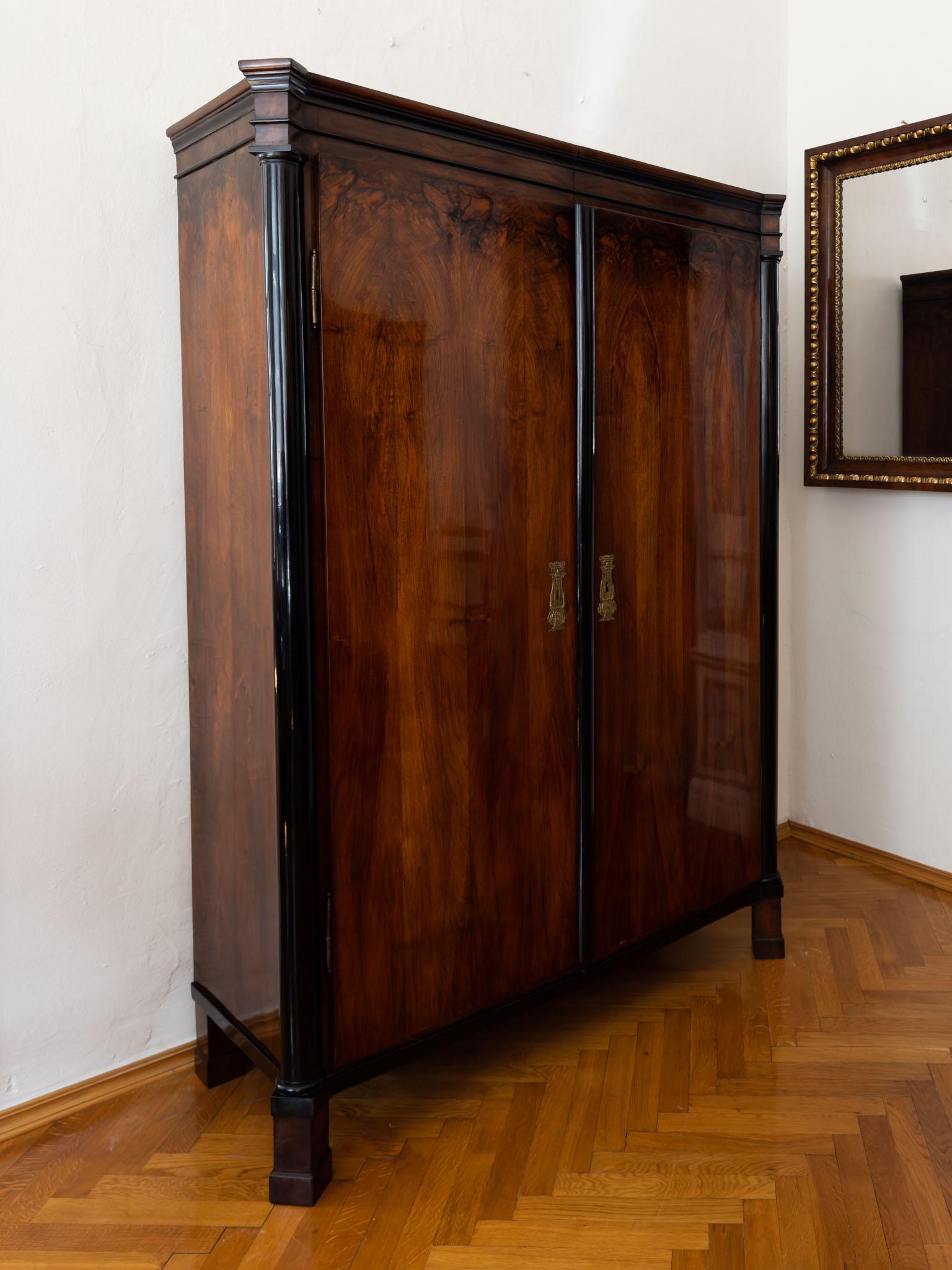 Two-door wardrobe with profiled cornice and ebonised columns. The corners of the wardrobe are cranked and rest on socketed square feet. Very beautiful walnut veneer on the front. The interior division consists of a clothes rail with one shelf.