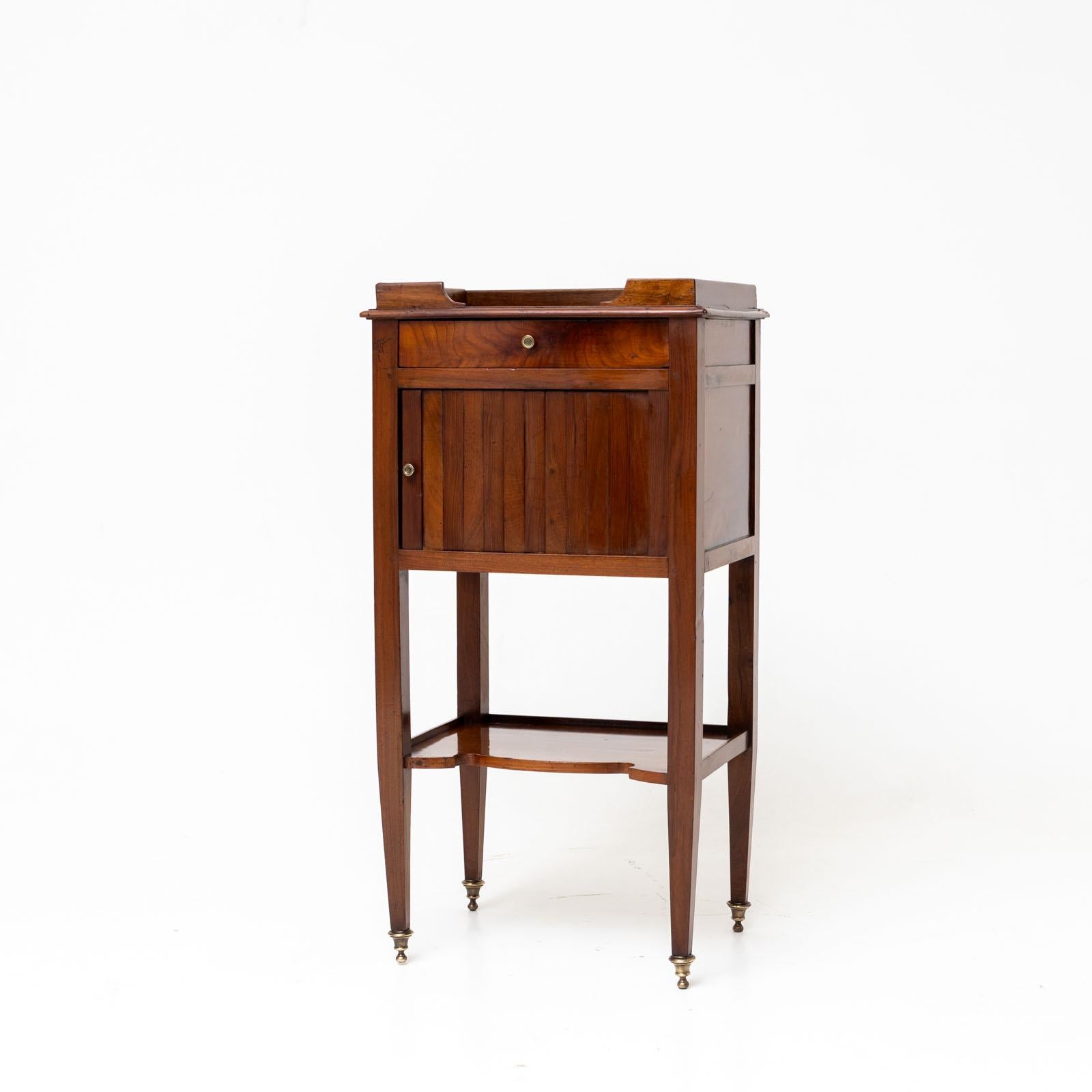 Small Biedermeier bedside cabinet with tambour door, small drawer and one shelf. The cabinet stands on brass sabots and the top is surrounded by a small bar. The bedside cabinet is veneered in walnut and polished by hand.