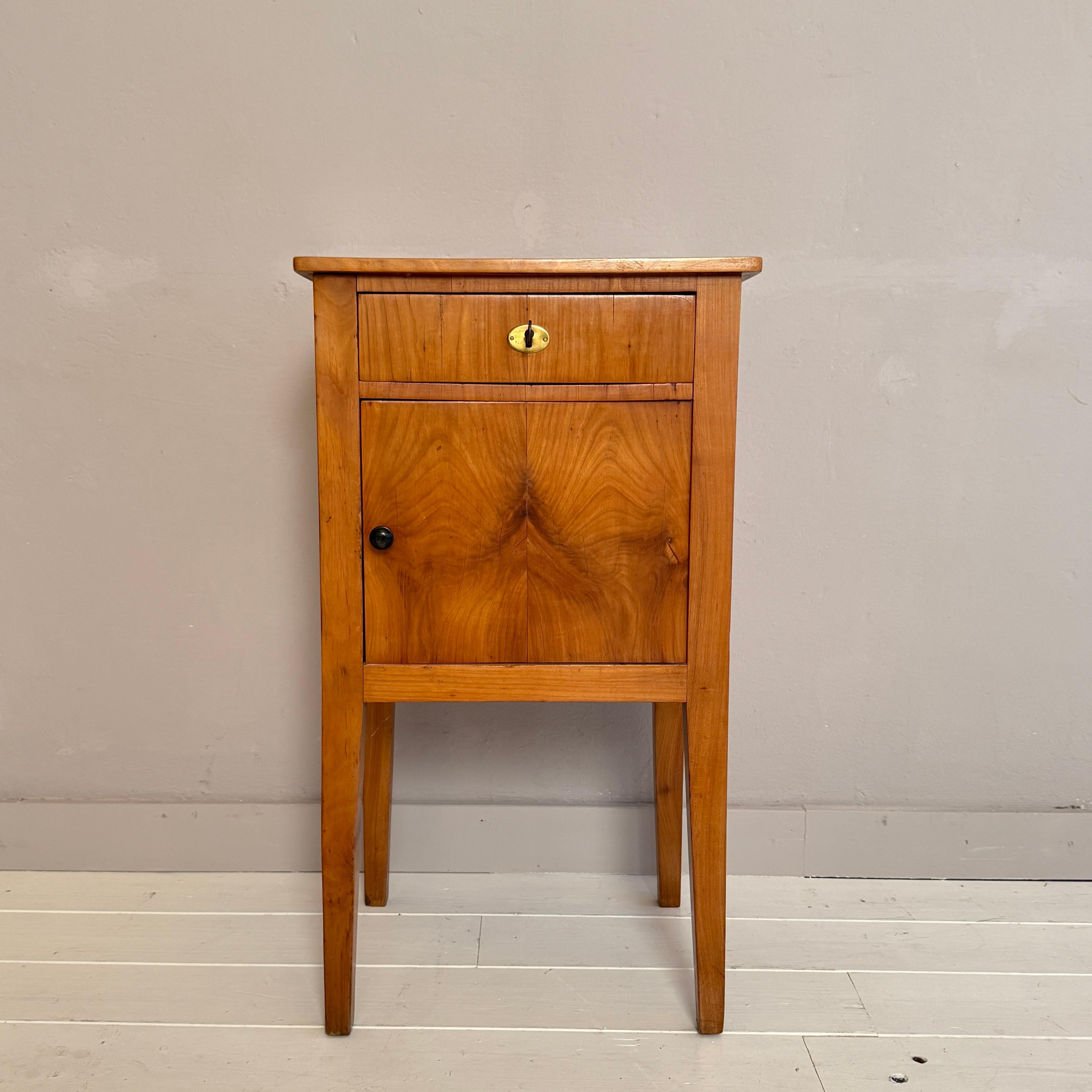 Questo affascinante comodino Biedermeier del XIX secolo, realizzato in ciliegio intorno al 1820, cattura la sobria eleganza e la calda bellezza naturale caratteristiche del periodo. Le linee pulite, le gambe affusolate e le proporzioni armoniose