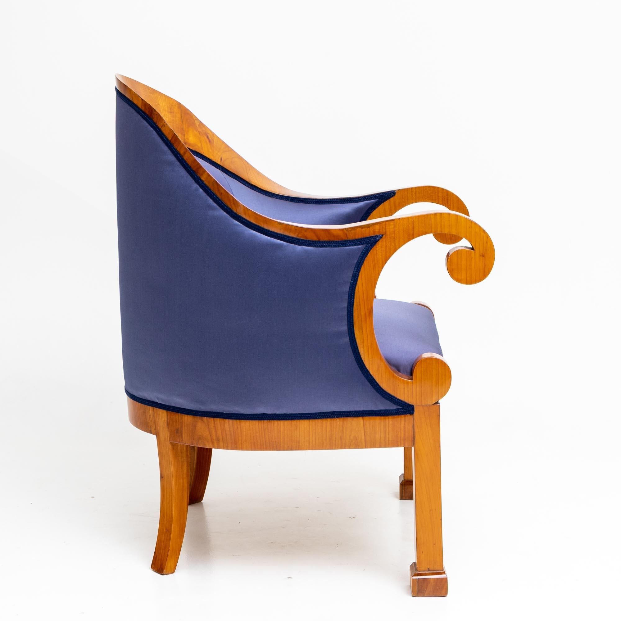 Biedermeier bergère in cherry veneer with elegant, c-shaped curved armrests and a centrally rising backrest. The armchair stands on socketed square feet at the front and flared sabre legs at the back. It is newly upholstered on all sides in a blue