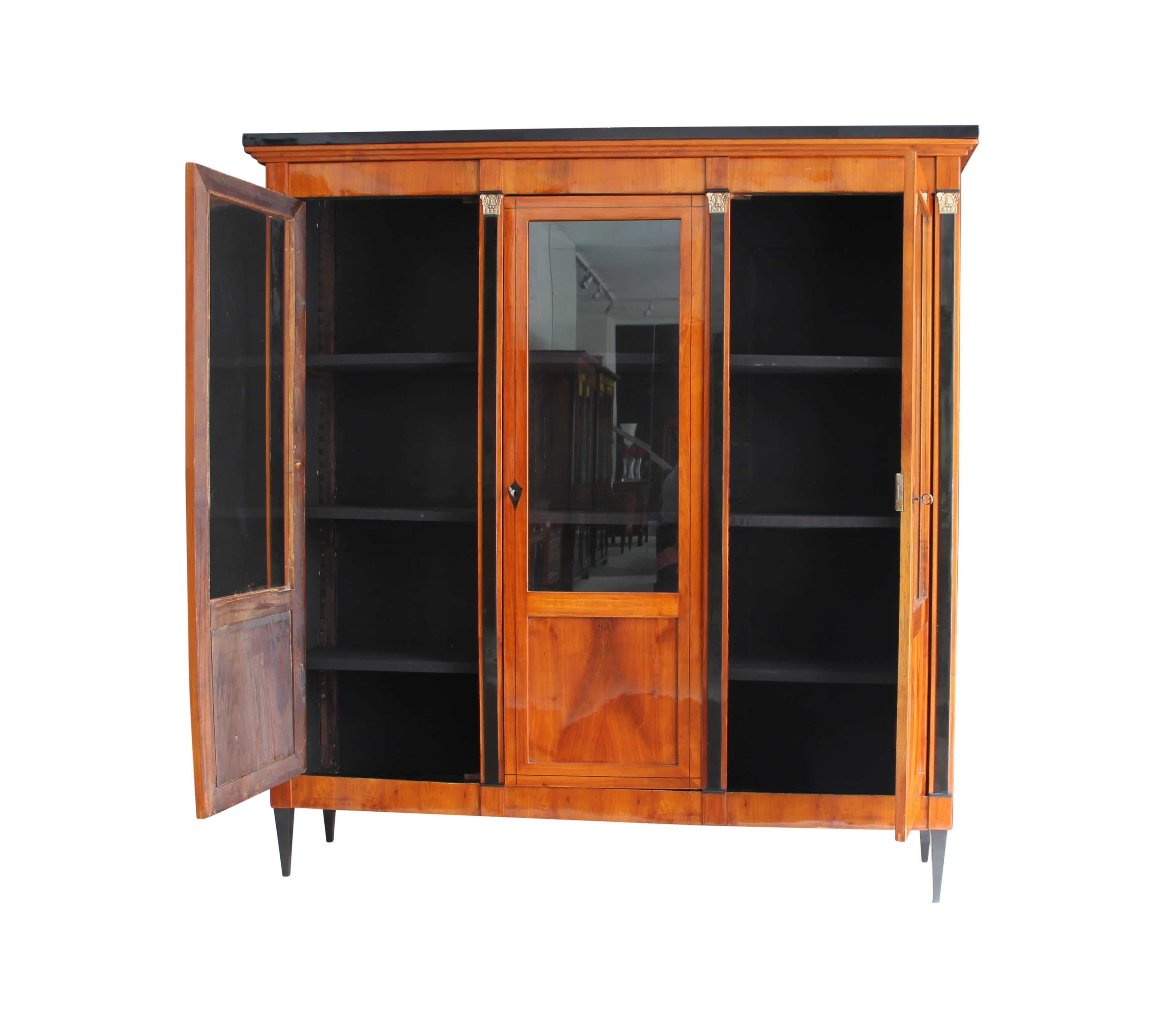 Very elegant three-doored Biedermeier bookcase from 2nd half of the 19th century.

Wonderful cherry veneer and solid wood. All original brass metals and old glasses.
The doors are separated with ebonized pilaster columns with brass capitals.

On the