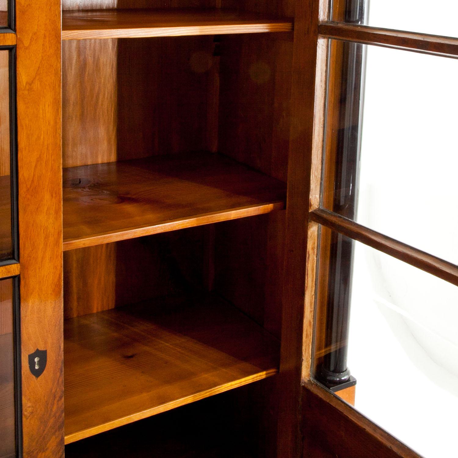 Two-door walnut Biedermeier bookcase with an angular pediment with ebonized moldings as well as ebonized columns. The doors show three-quarter glazing and have fillings with ebonized moldings. The bookcase is in a restored condition.
