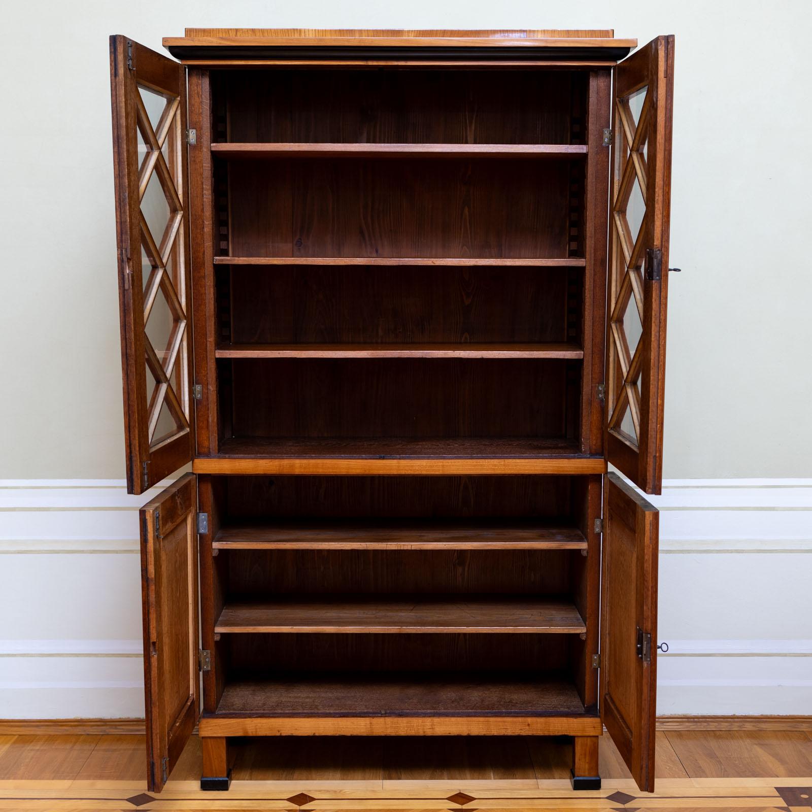 Biedermeier Bookcase with a two-door base and a two-door top with glazed doors with rhomboid strutting. The cabinet ist veneered in cherry and stands on square feet with ebonised details. The cabinet has been restored and polished by hand. 