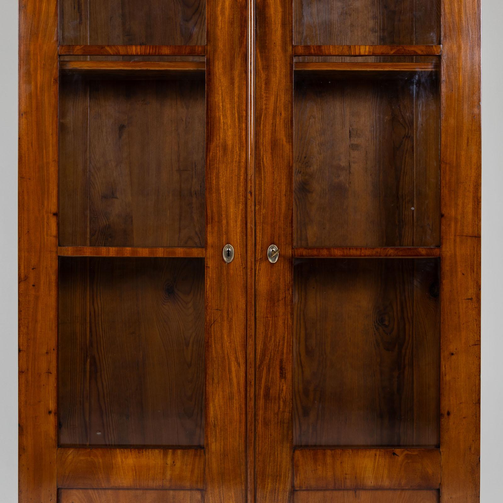 Biedermeier bookcase with two doors with three-quarter glazing and a flat profiled cornice. The cabinet stands on square feet and is veneered in mahogany. The interior is fitted with four adjustable shelves. The bookcase has been restored and