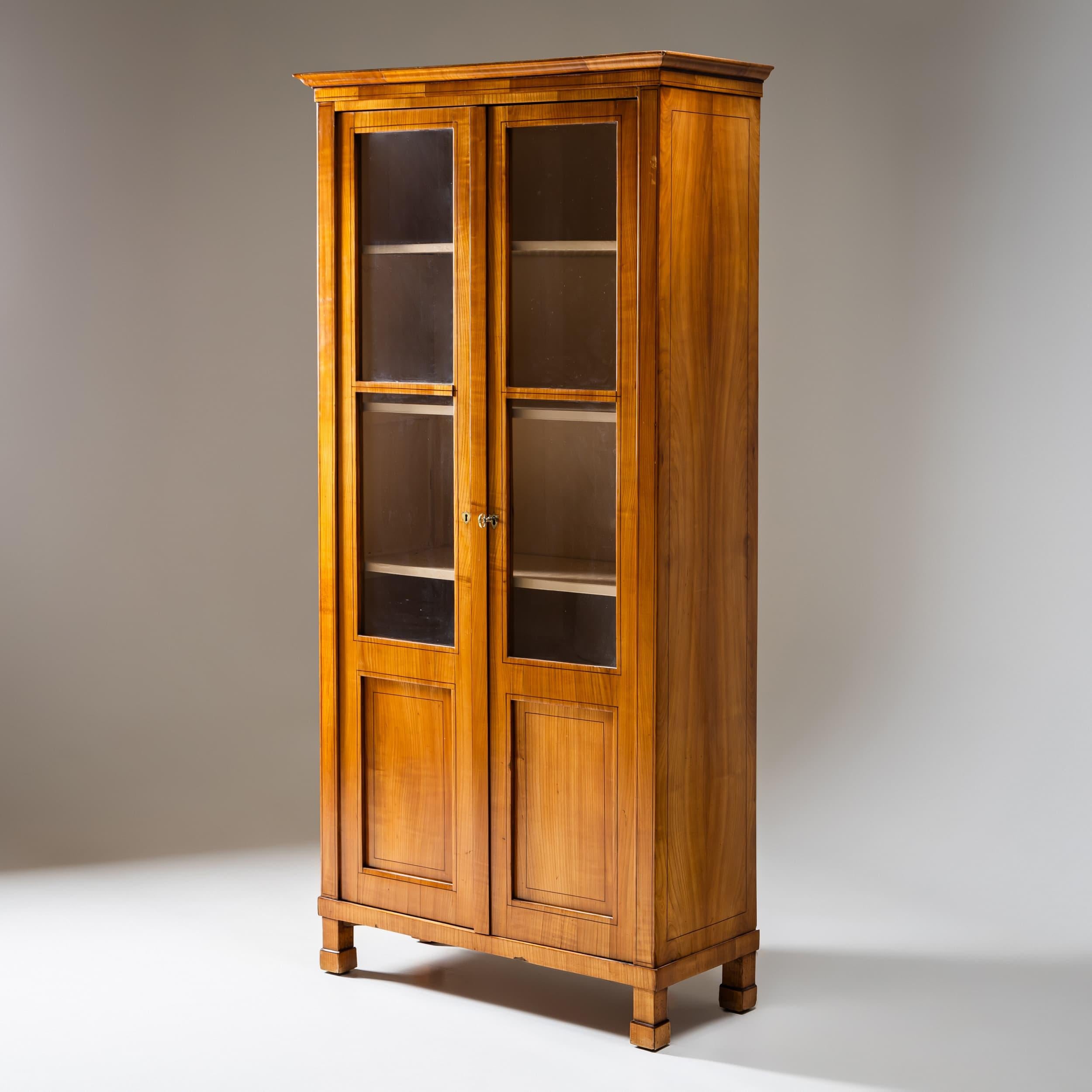 Two-door bookcase from the Biedermeier period, dating to around 1820. The doors are two-thirds glazed, allowing for an elegant display of books or collectibles.
The bookcase stands on plinth-mounted square legs and is veneered in cherrywood. The