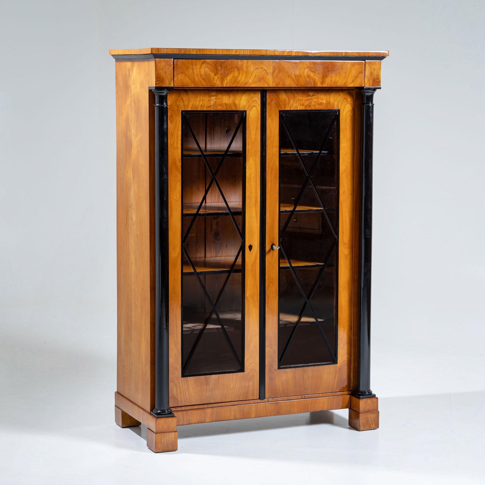 Libreria in ciliegio con colonne laterali ebanizzate e reticolo a forma di diamante sulle ante vetrate. All'interno, il mobile offre molto spazio con quattro ripiani con bordi frontali ebanizzati che creano un accento di stile. Inoltre, il mobile è