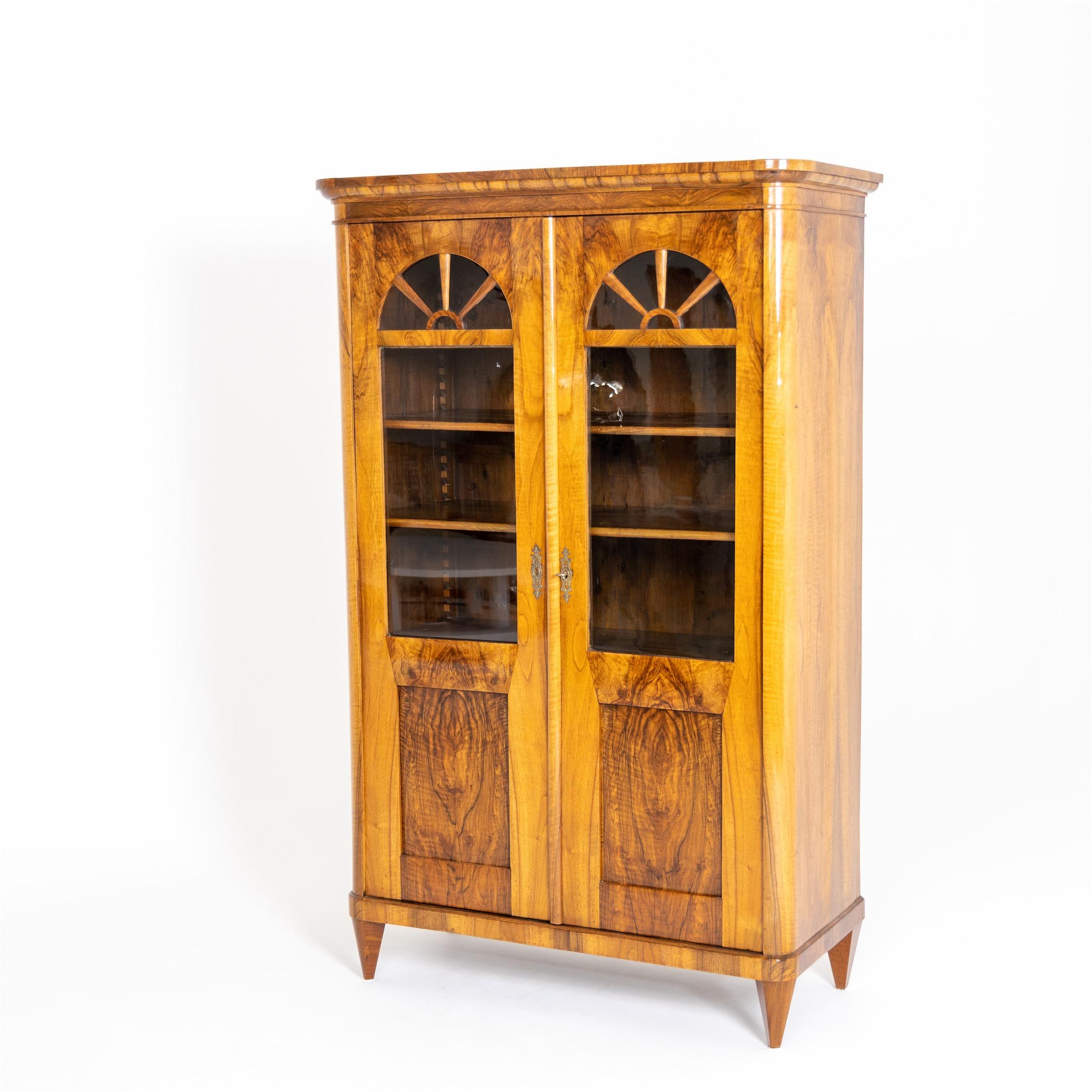 Two-door bookcase on conical legs with rounded corners and two-thirds glazing of the doors, which ends in a segmental arch in the upper area. The interior consists of shelves that can be adjusted in height. Very beautiful walnut veneer, hand