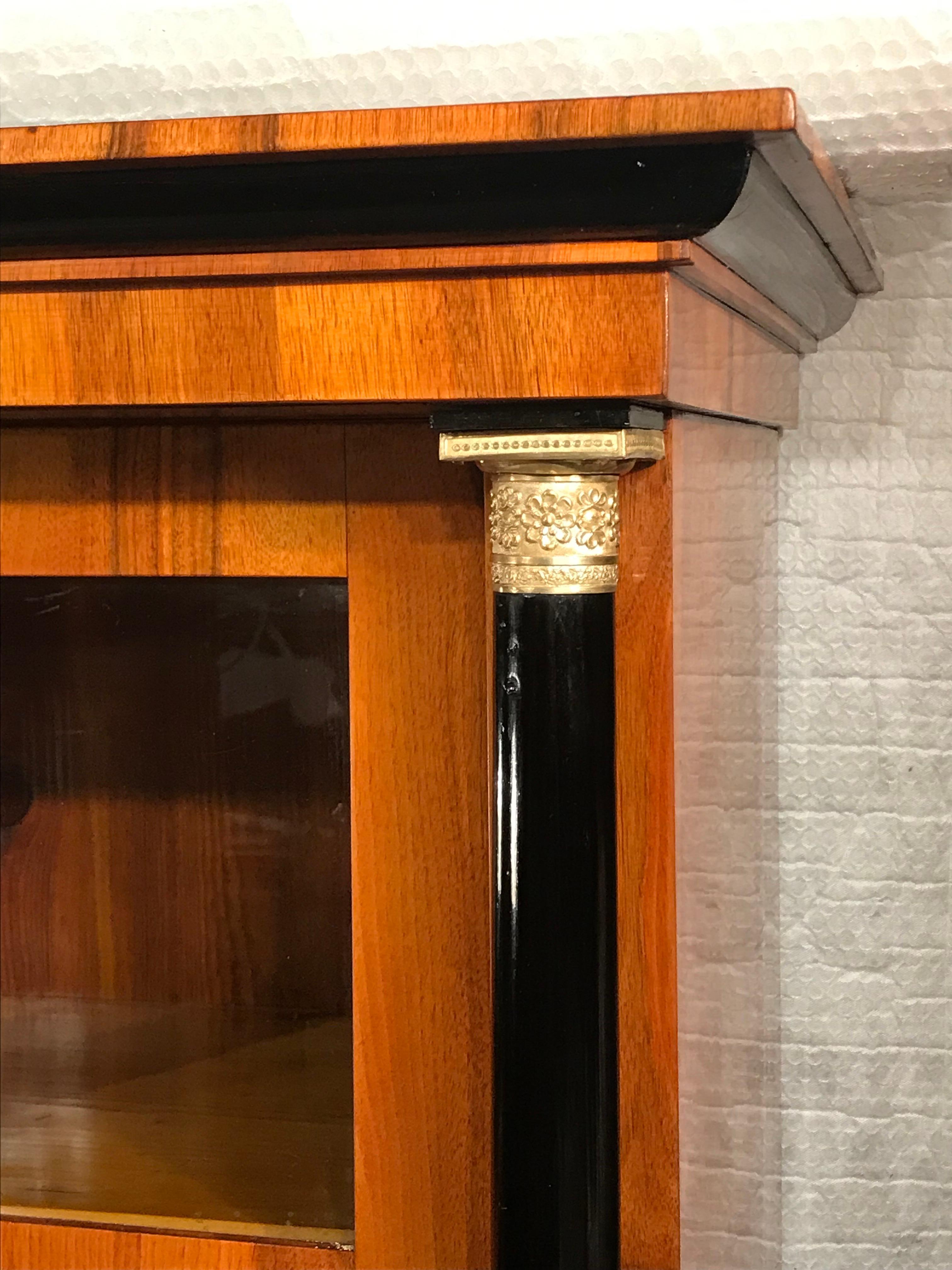 This unique Biedermeier bookcase dates back to around 1820 and comes from Southern Germany. the bookcase has a pretty walnut veneer with two transome glass doors. They are framed by two ebonized columns with original repousse brass capitals and
