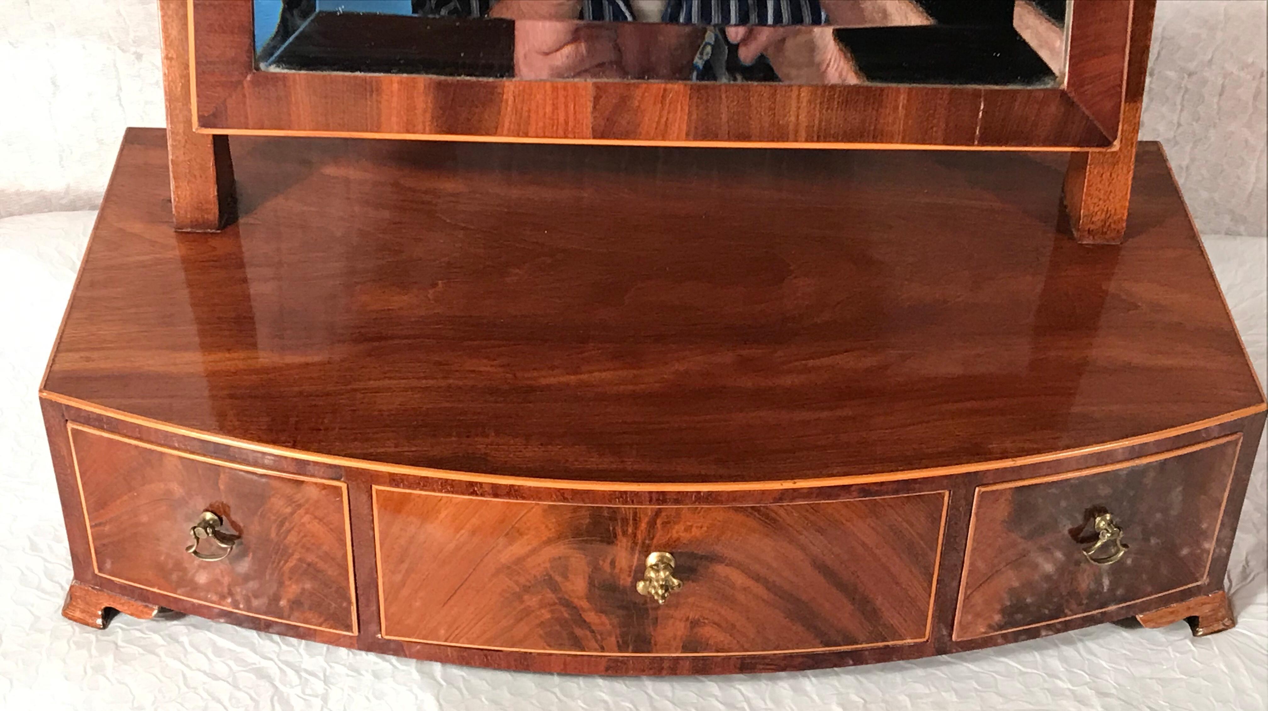 This Biedermeier bow front dressing table mirror has three drawers. It is embellished with mahogany veneer and ash inlays. The mirror has its original mirror glass. It dates back to around 1825 and comes from Berlin in Germany. It is a very elegant