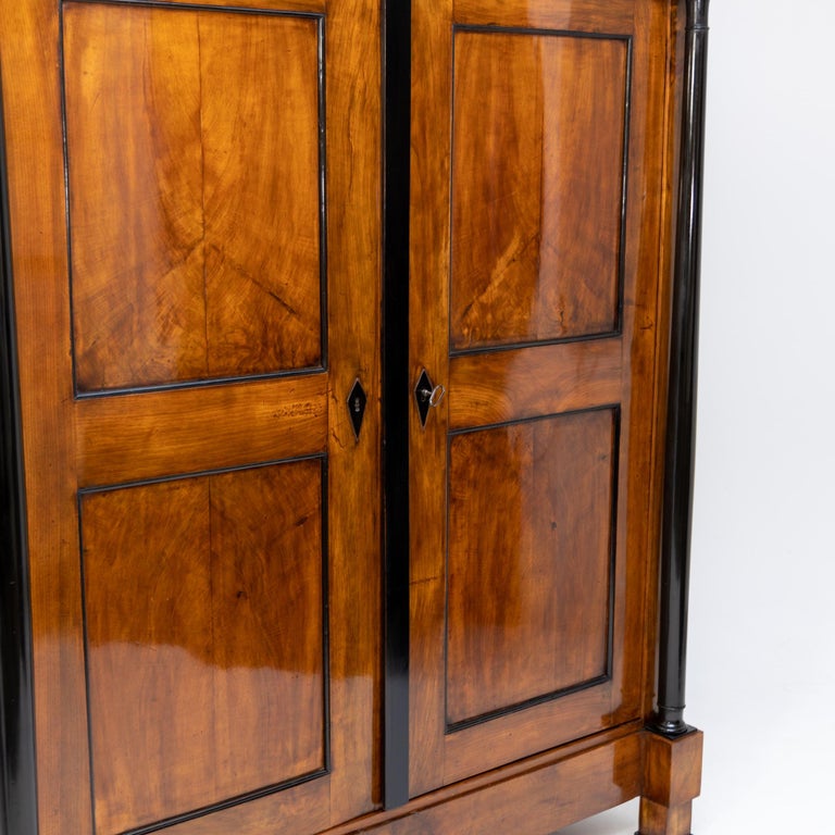 Two-door Biedermeier wardrobe in walnut veneer with ebonised columns and profiles. The interior consists of a hat shelf and a clothes rail. The wardrobe has been professionally refurbished and hand polished.