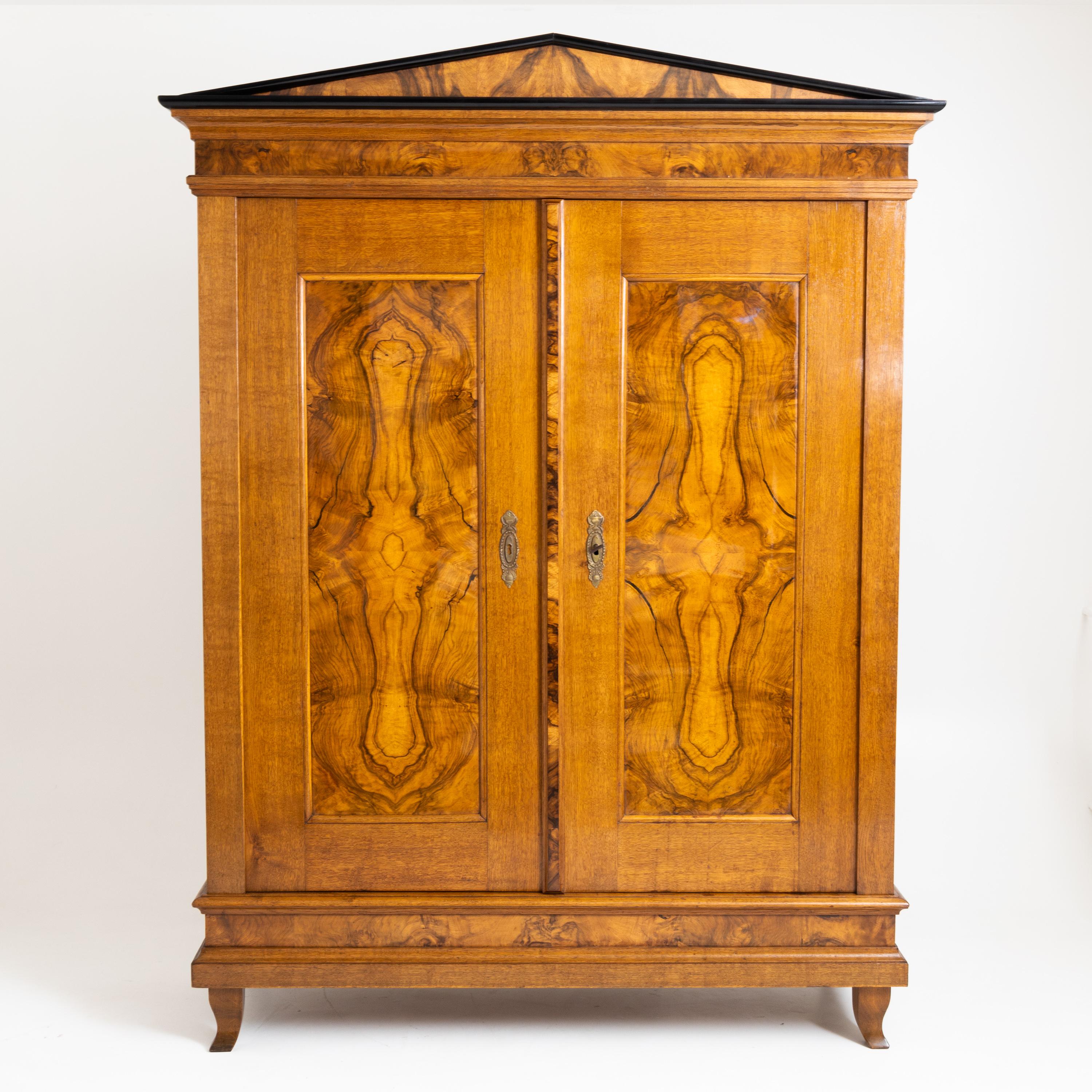 Large two-door wardrobe with walnut-veneered panels and oak body. The cornice in classicist design with ebonized mouldings around the tympanum field. The flap moulding is also veneered in walnut on the top. The sides of the body are coffered like