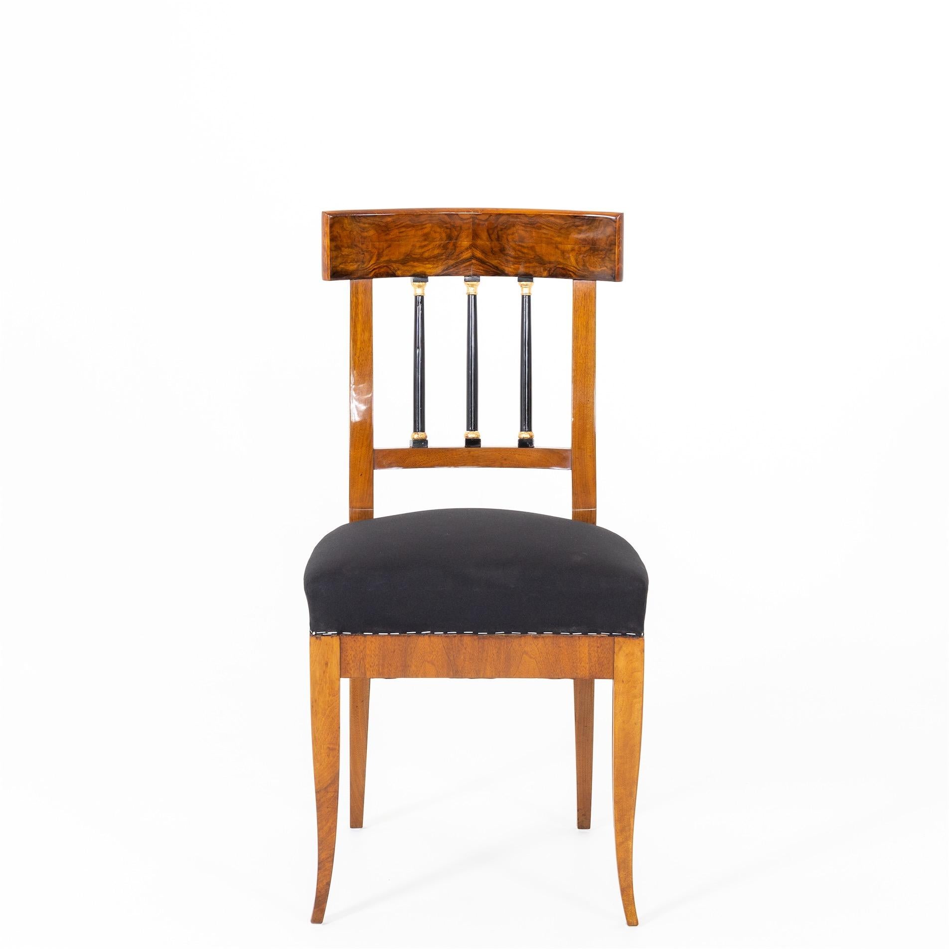 Biedermeier chair made of solid and veneered walnut. The chair stands on elegantly curved tapered legs and the back features ebonized solid columns with gold-patinated capitals. The chair is covered with a black base fabric.
 