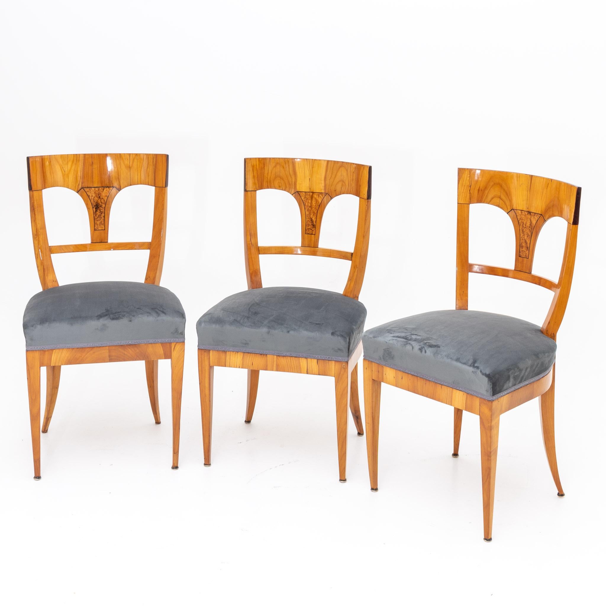 Set of three Biedermeier chairs in cherry with trapezoidal openwork backrest with straight top rail and center splat with inlaid birch burl veneer. The seats are reupholstered with a gray-blue velvet fabric.