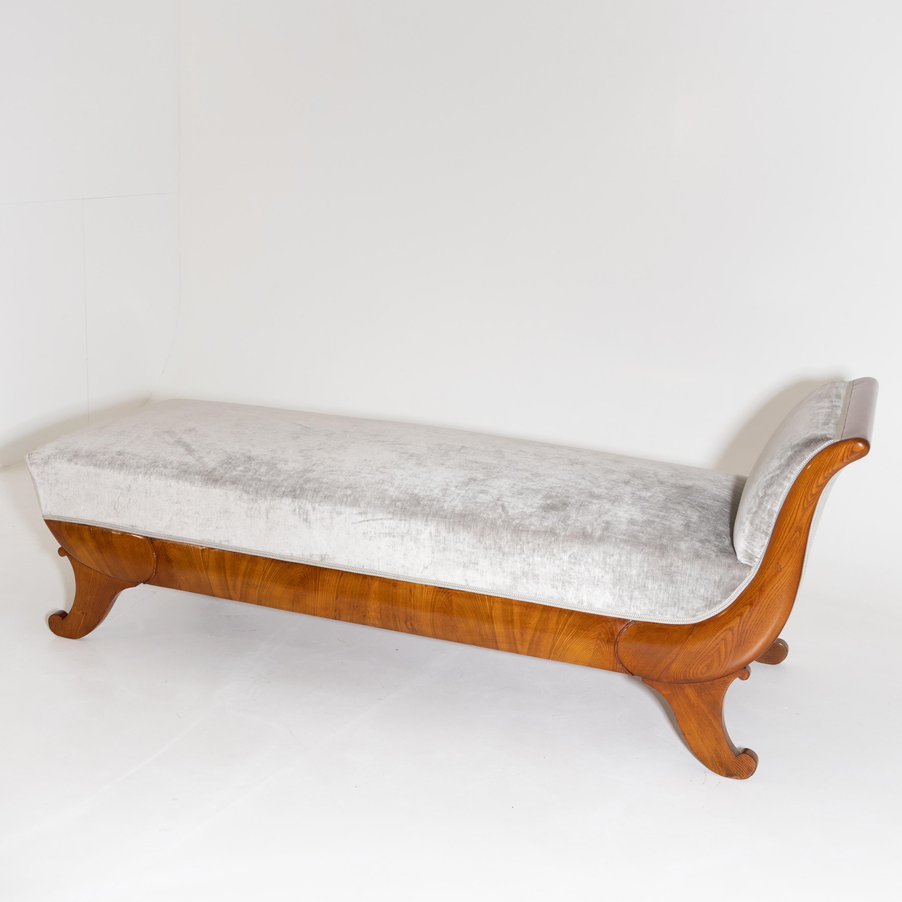 Biedermeier recamiere in ash, with sledge-shaped backrest standing on stylized volute feet. The straight frame is slightly rounded and hand polished. The recamiere has been newly upholstered on all sides with a smoky blue velvet fabric.