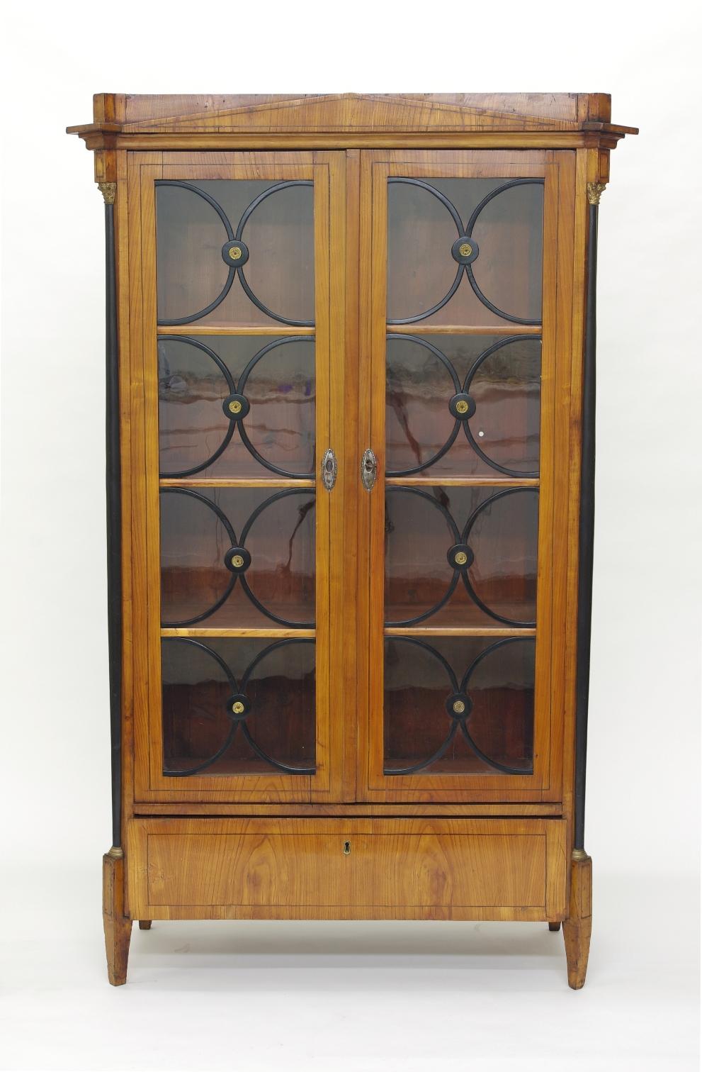 Biedermeier cherry bookcase, circa 1820. Biedermeier cherry bookcase/vitrine, the top with a projecting pediment over two doors, each with ebonized mullions and brass medallions opening to three adjustable shelves, the canted sides with ebonized